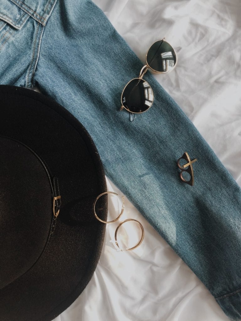 An OOTD style flatlay on a white sheet, featuring a denim jacket, gold accessories and a black hat