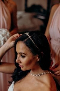 Bethany adjusting her headband in the mirror while her bridesmaid fastens her necklace