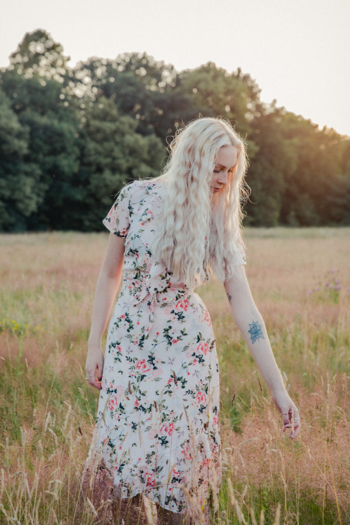 Amber wearing a pale pink floral dress, stood touching the tall grass at sunset during a golden hour photoshoot