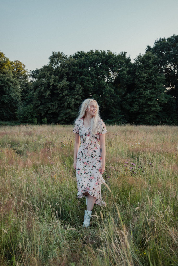 Amber wearing a pale pink floral dress, walking through the tall grass at sunset during a golden hour photoshoot