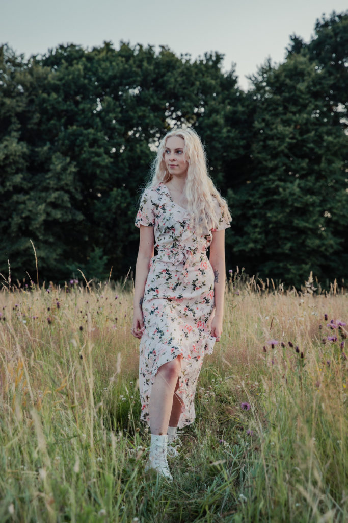 Amber wearing a pale pink floral dress, stood in the tall grass at sunset during a golden hour photoshoot