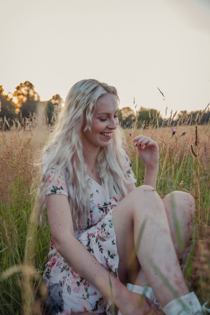 Amber wearing a pale pink floral dress, sat in the tall grass at sunset during a golden hour photoshoot