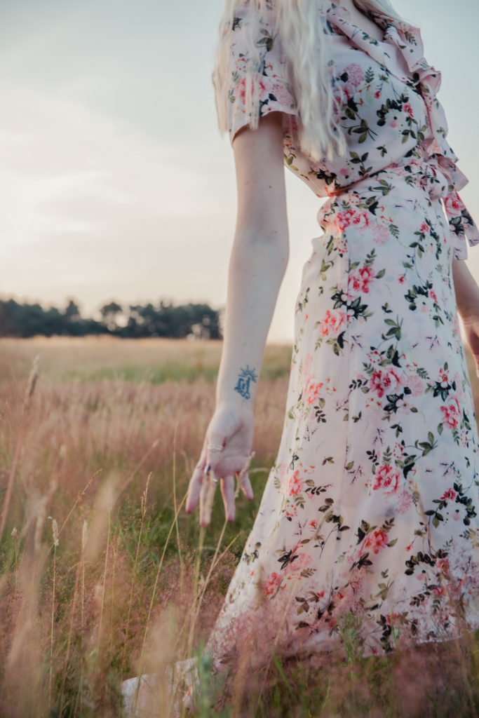 Amber wearing a pale pink floral dress, walking through and touching the tall grass at sunset during golden hour