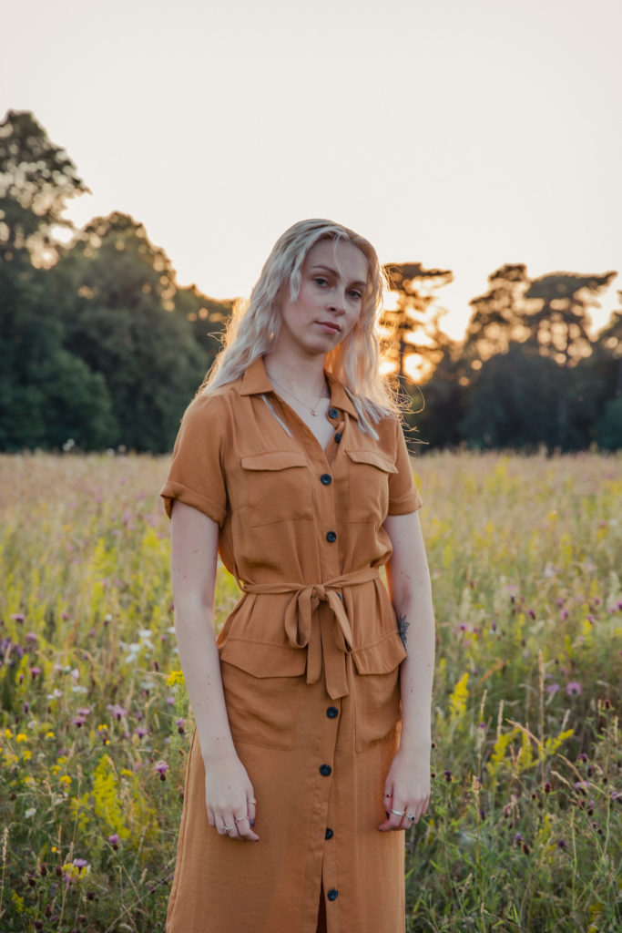 Amber wearing a button up burnt orange dress, stood in the tall grass at sunset during a golden hour photoshoot