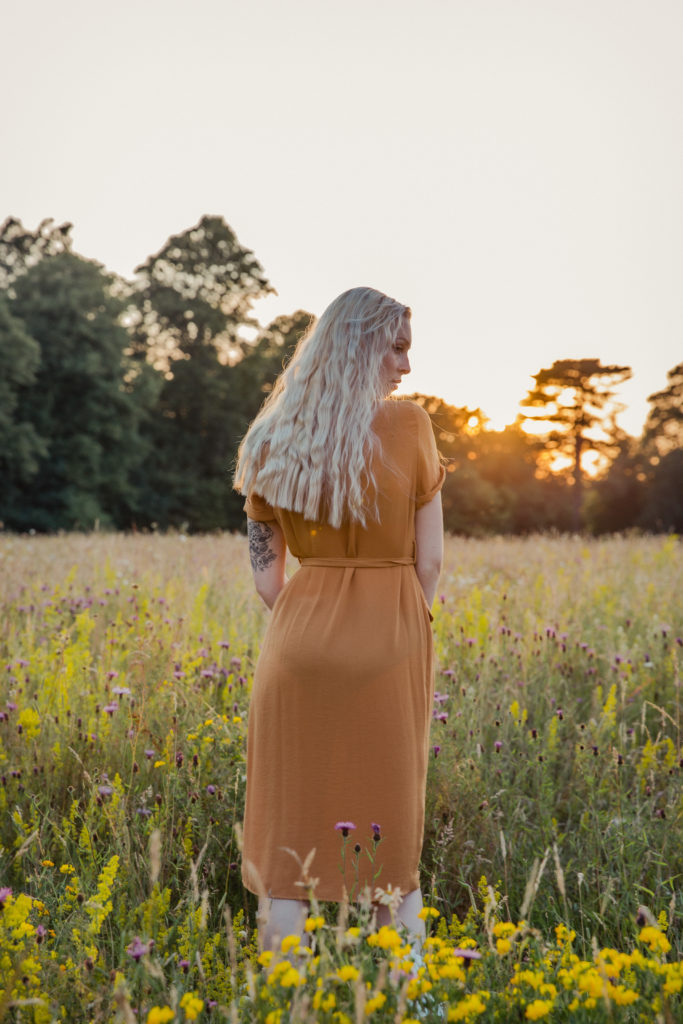 Amber wearing a button up burnt orange dress with her back to the camera, stood in the tall grass at sunset