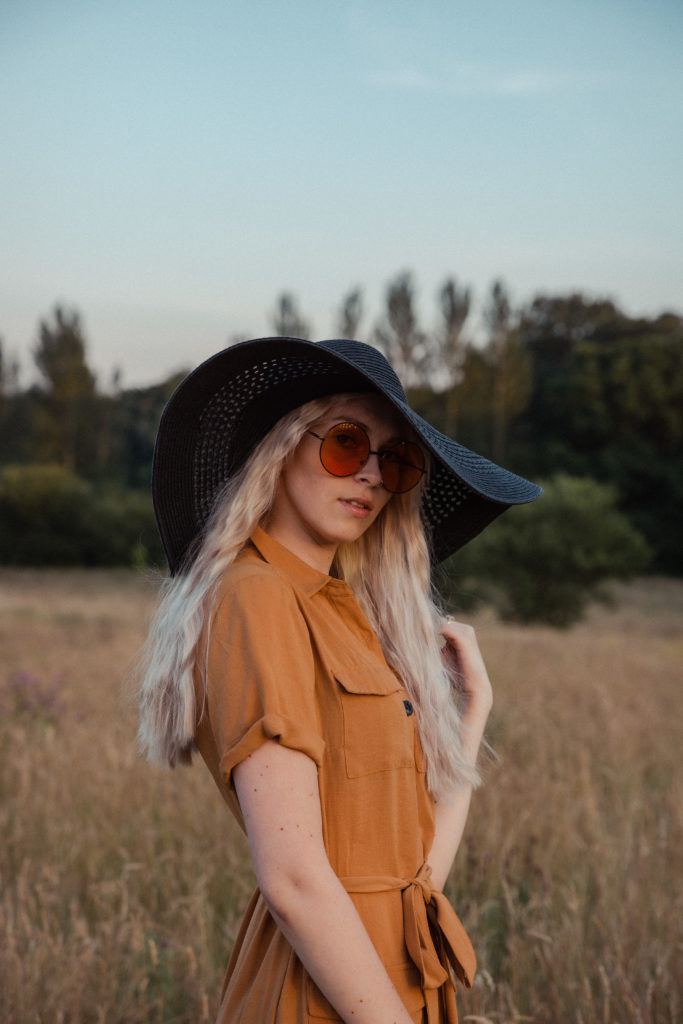 A close up of Amber wearing a button up burnt orange dress with a black straw hat and round orange sunglasses, stood in the tall grass at sunset during a golden hour photoshoot