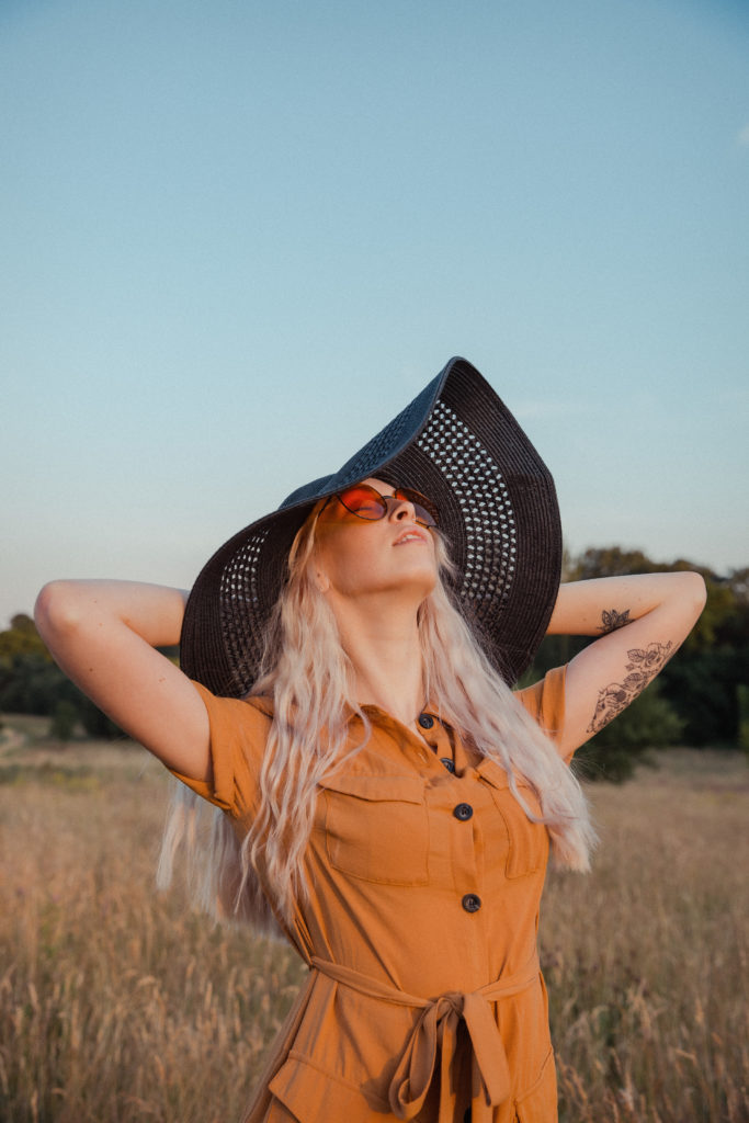 A close up of Amber wearing a button up burnt orange dress with a black straw hat and round orange sunglasses, stood in the tall grass with her head back, facing the sun at sunset
