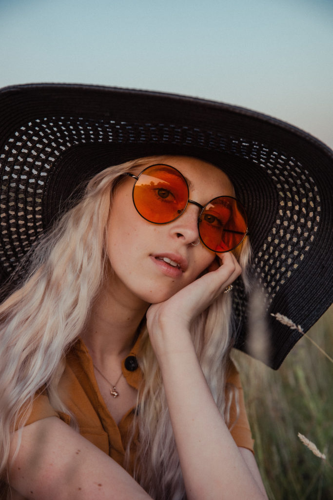 A close up of Amber wearing a button up burnt orange dress with a black straw hat and round orange sunglasses, sat in the tall grass at sunset during golden hour