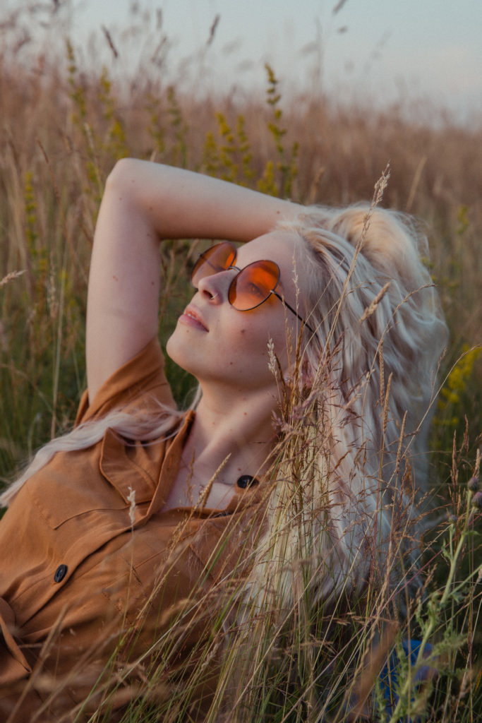 A close up of Amber wearing a button up burnt orange dress with round orange sunglasses, laying in the tall grass, running her hands through her blonde hair at sunset