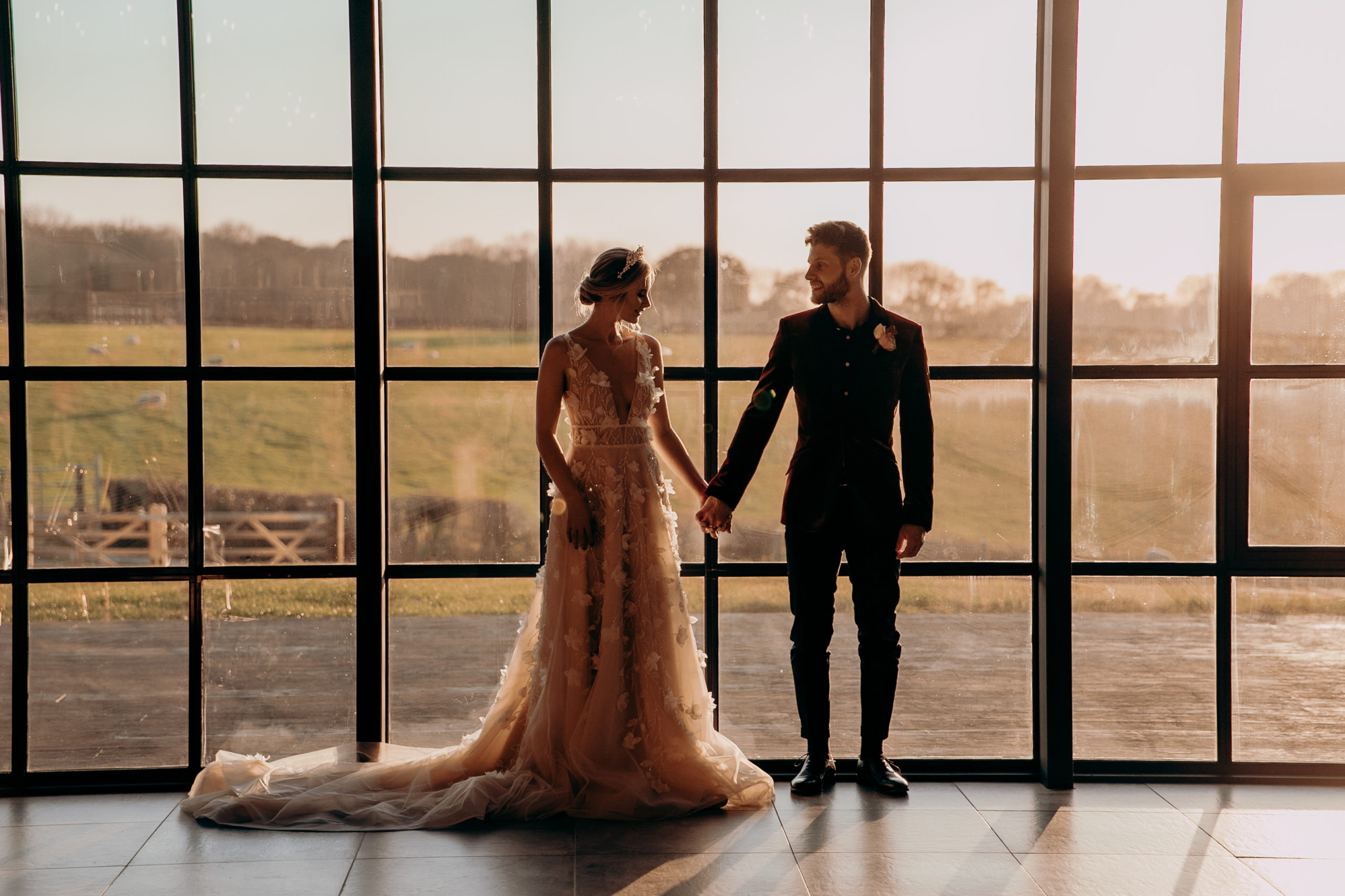 Zac and Esme stood holding hands against tall windows at sunset at Botley Hill Barn