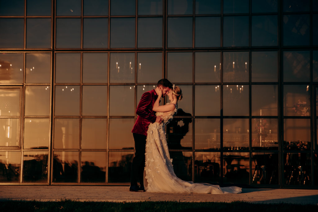 Zac and Esme embrace in their wedding outfits outside on the deck of Botley Hill Barn, infront of the windows at sunset