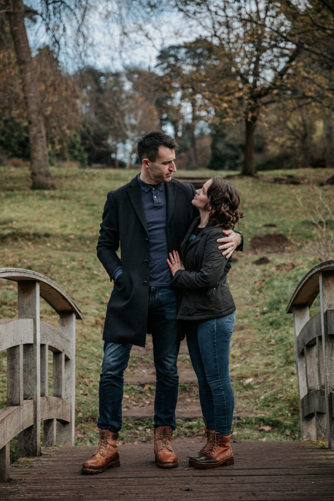 Laura and Stuart stand and cuddle on a little wooden bridge during their winter engagement shoot