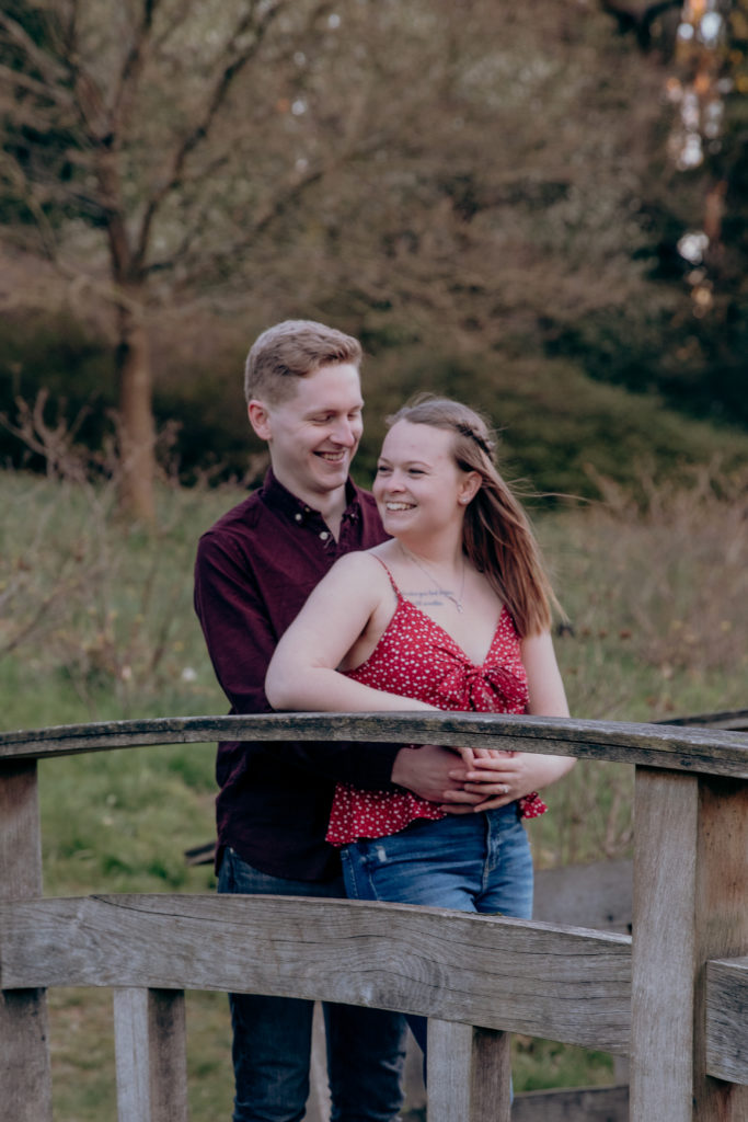 Toby and Jemma smile and laugh on a small wooden bridge during their spring engagement shoot