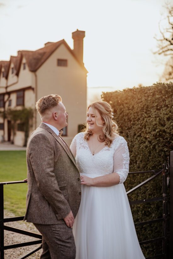 Bride and groom stand infront of a metal gate at sunset, with the Ufton Court Tudor house in the background