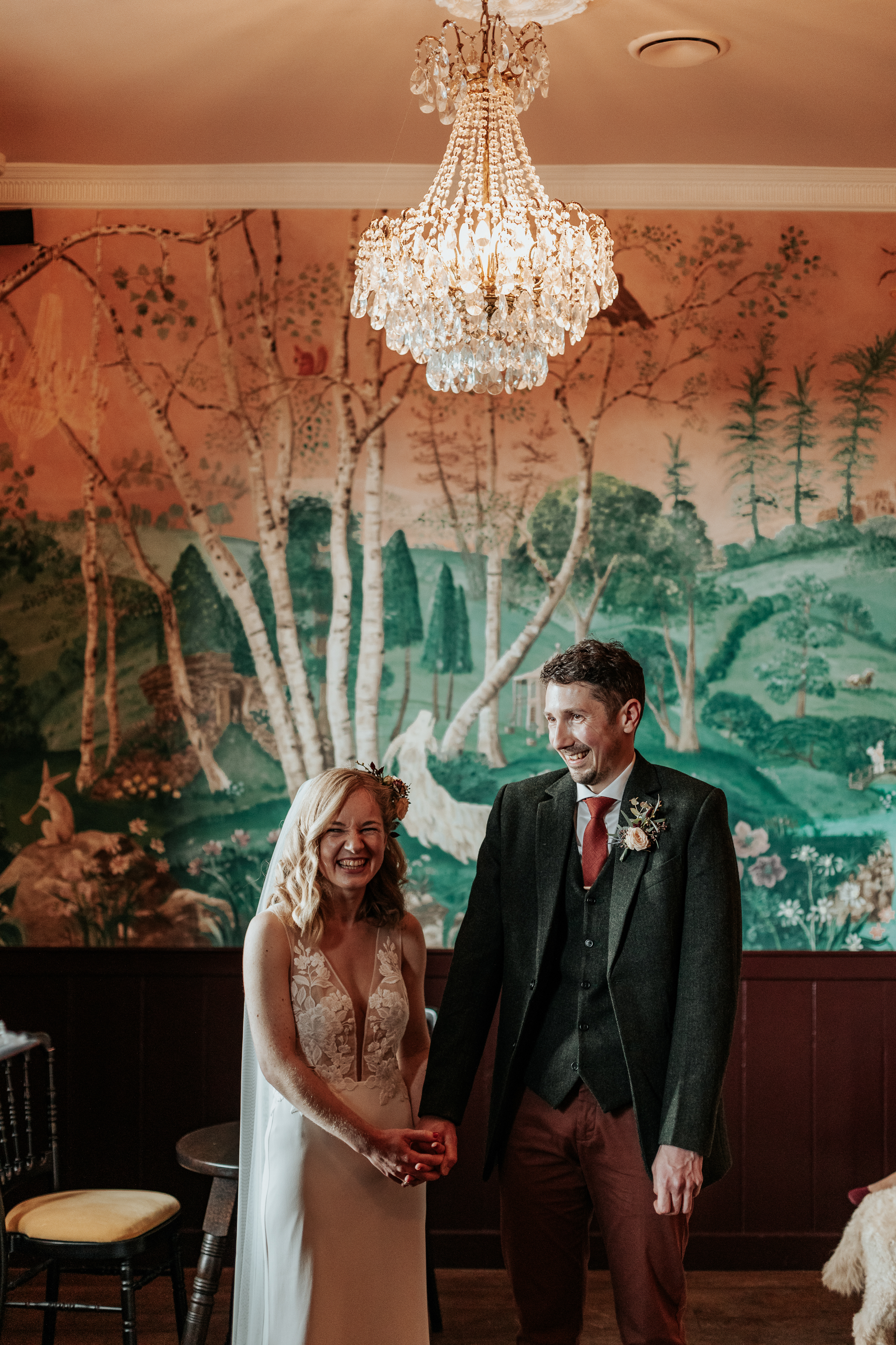 Maddie and Will stand holding hands below a chandelier during their wedding ceremony