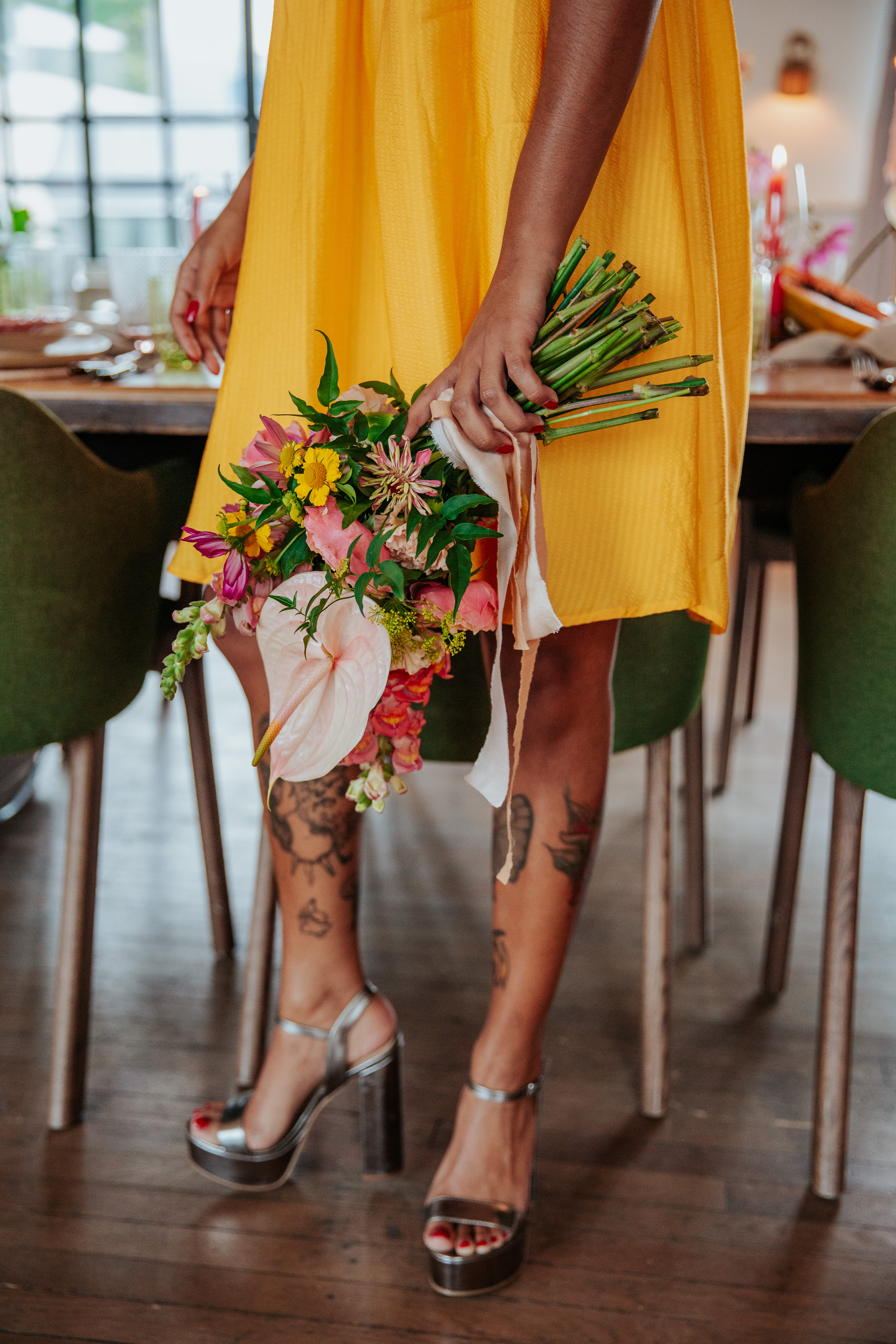 Meera holds her bouquet in a relaxed, downwards way with her yellow minidress and silver platform heels in the background