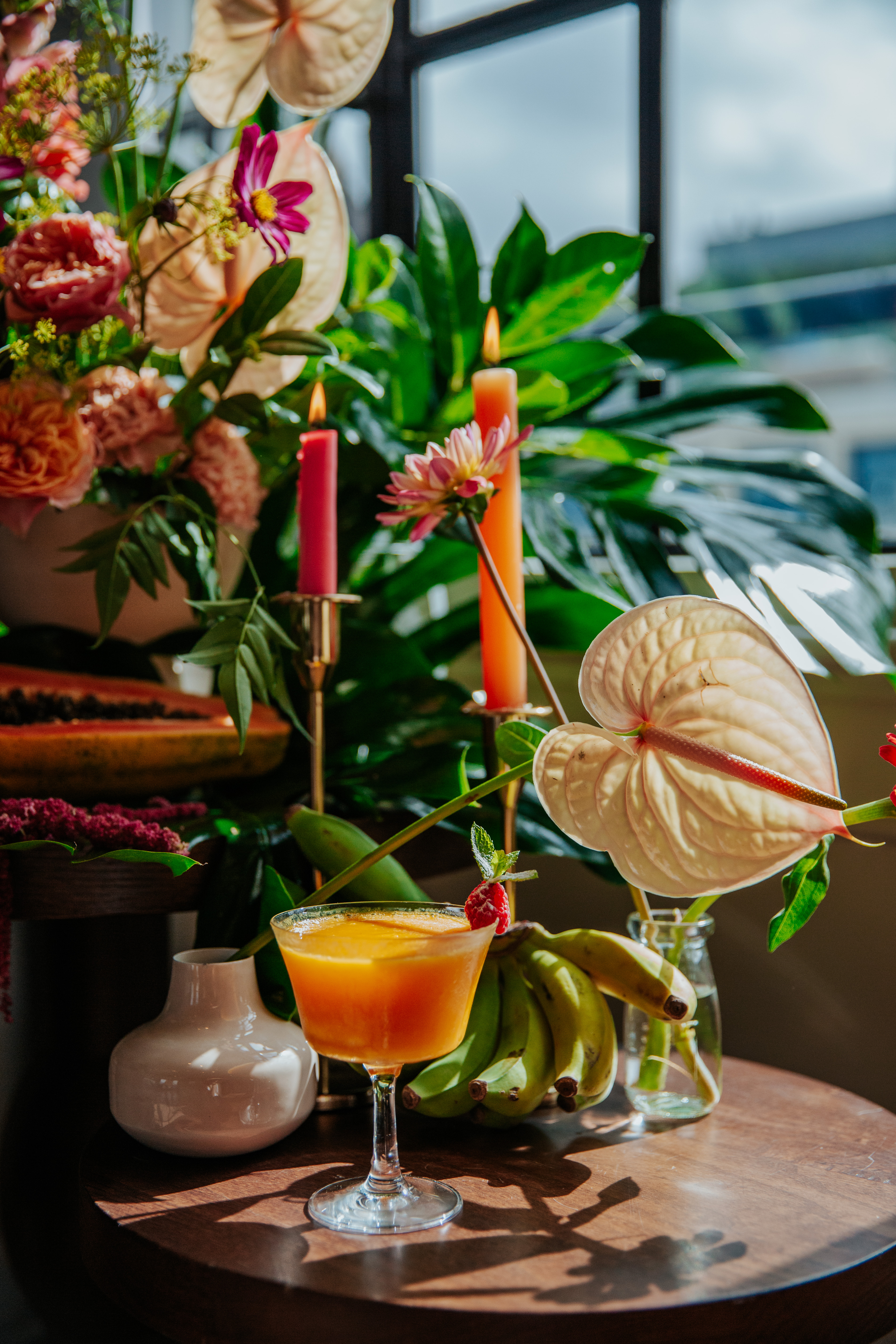 A close up shot of a floral display by Mortimer House's gallery windows, with leaves, candles, fruit and an orange cocktail