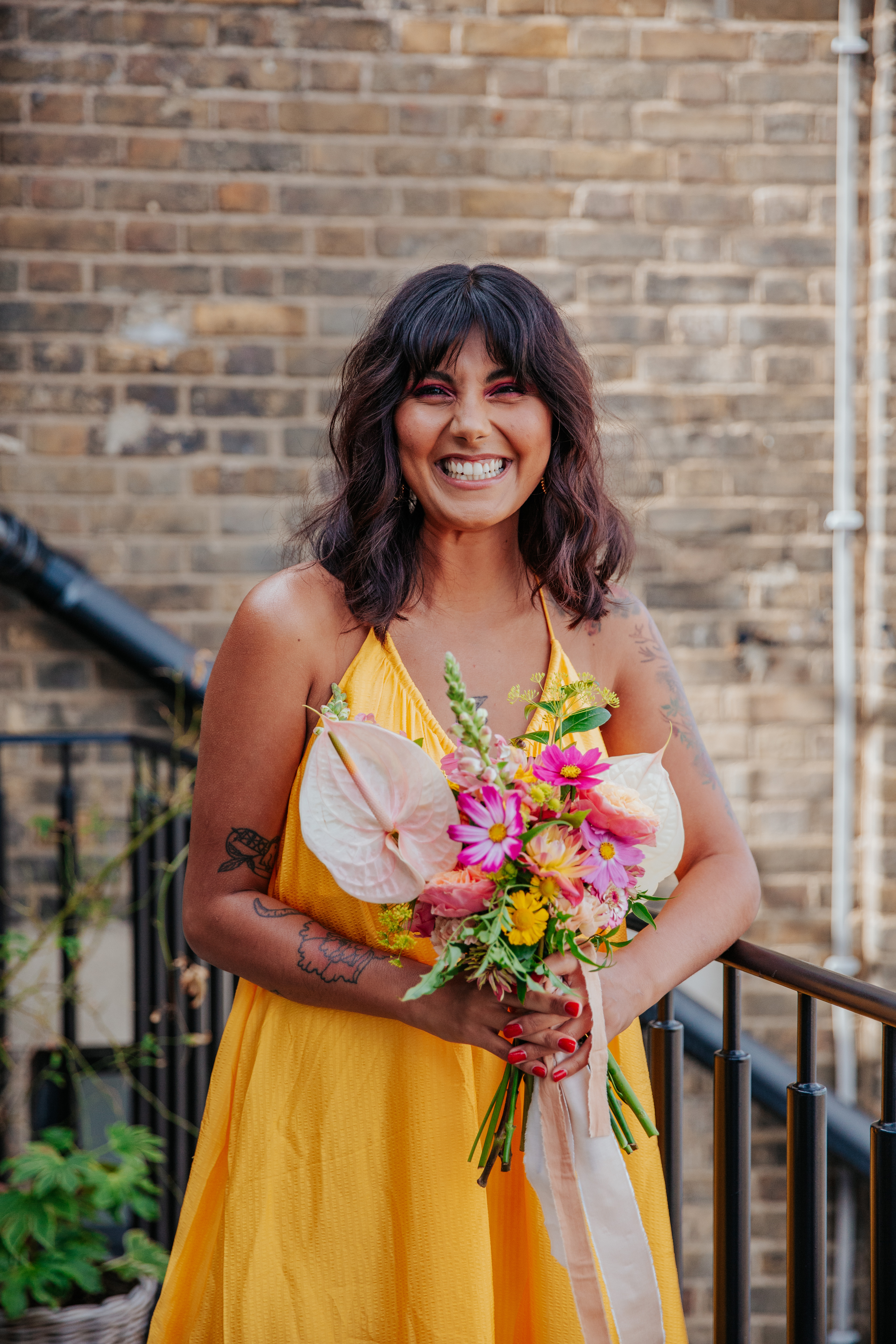 Meera leans against the balcony of Mortimer House while holding her bouquet and smiling