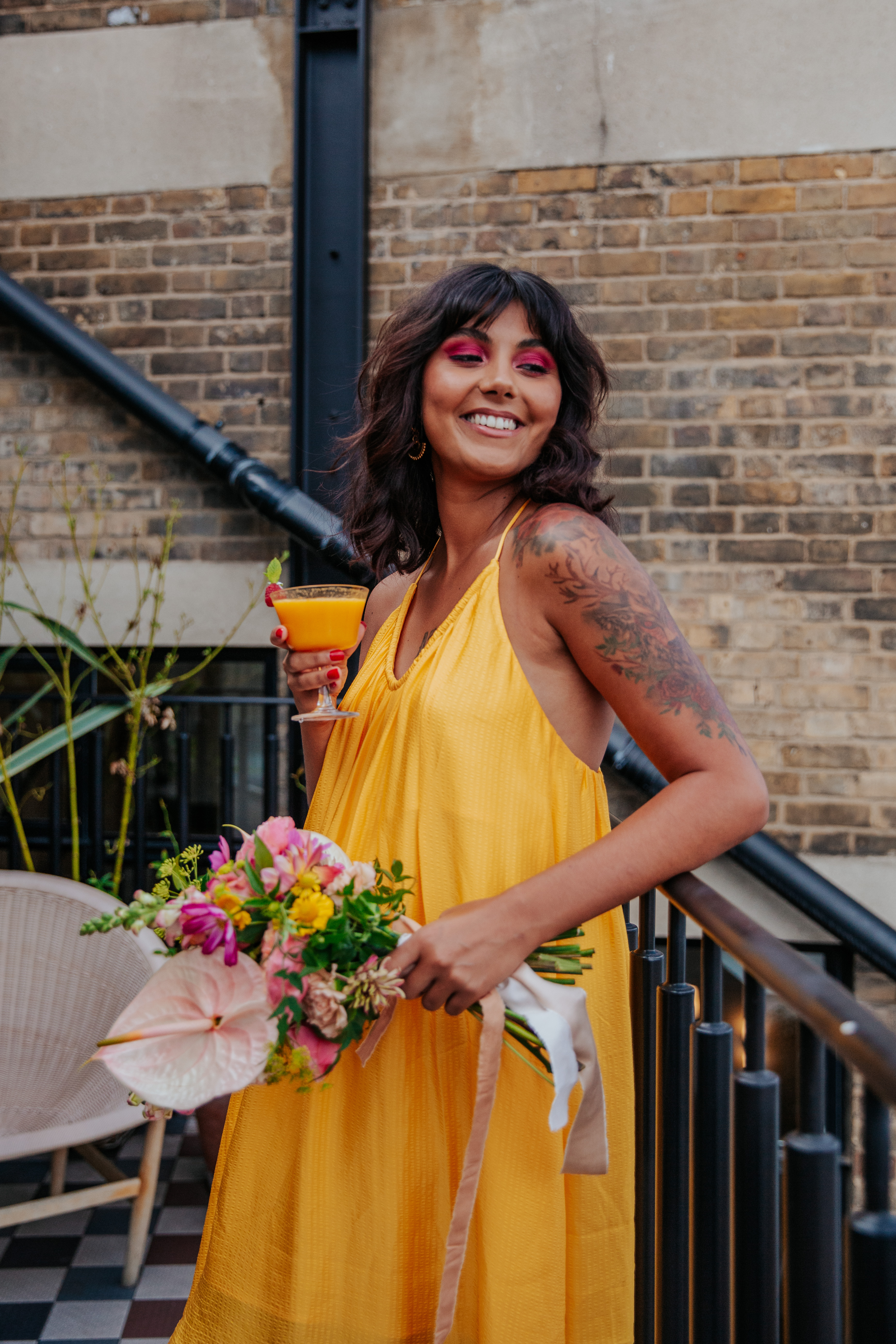 Meera smiles while leaning against the balcony of Mortimer House while holding her cocktial and bouquet