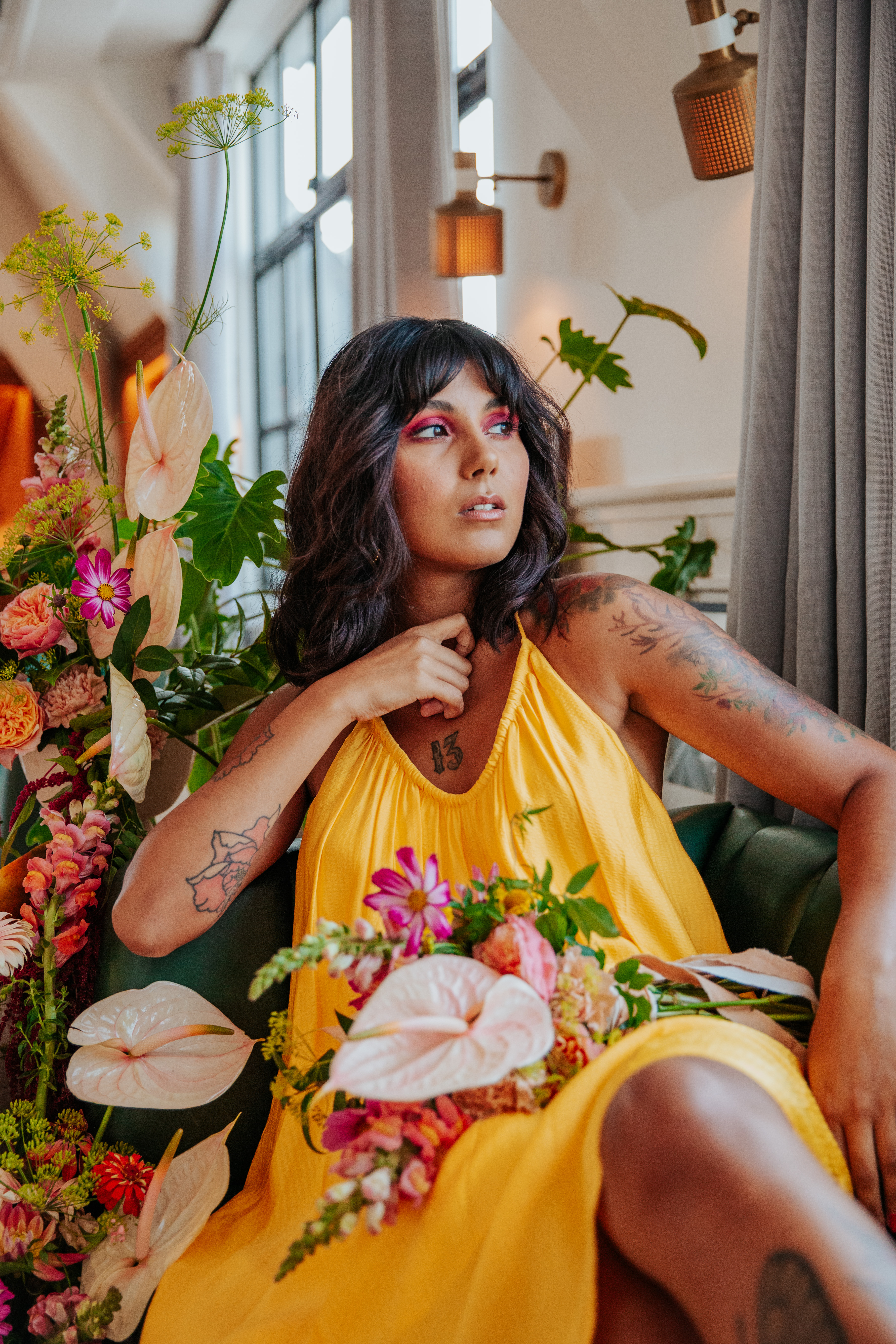 Meera lounges on a green leather booth, holding her bouquet next to a tropical floral display