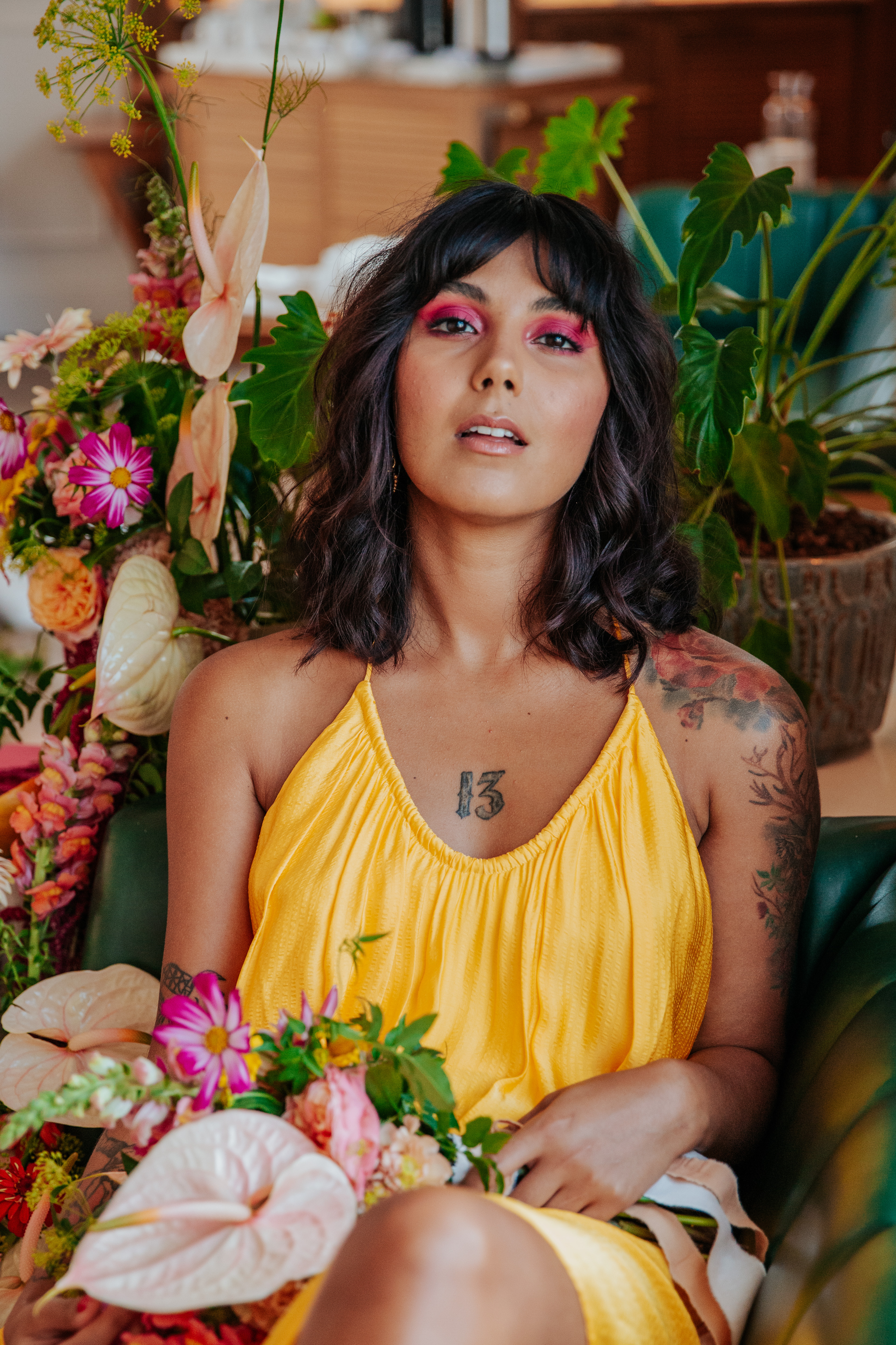 Meera lounges on a green leather booth, holding her bouquet next to a tropical floral display