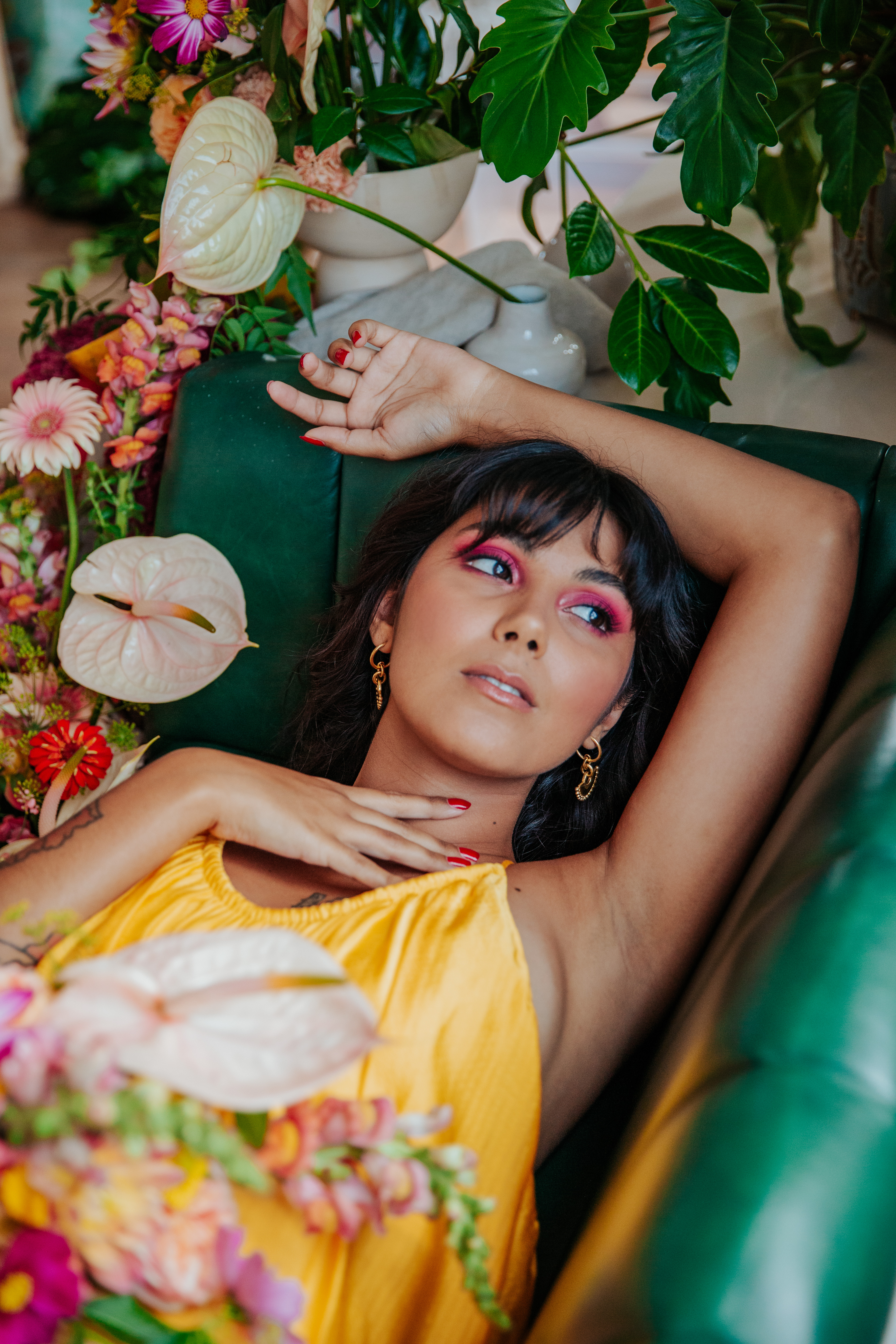 Meera lounges on a green leather booth, holding her bouquet next to a tropical floral display