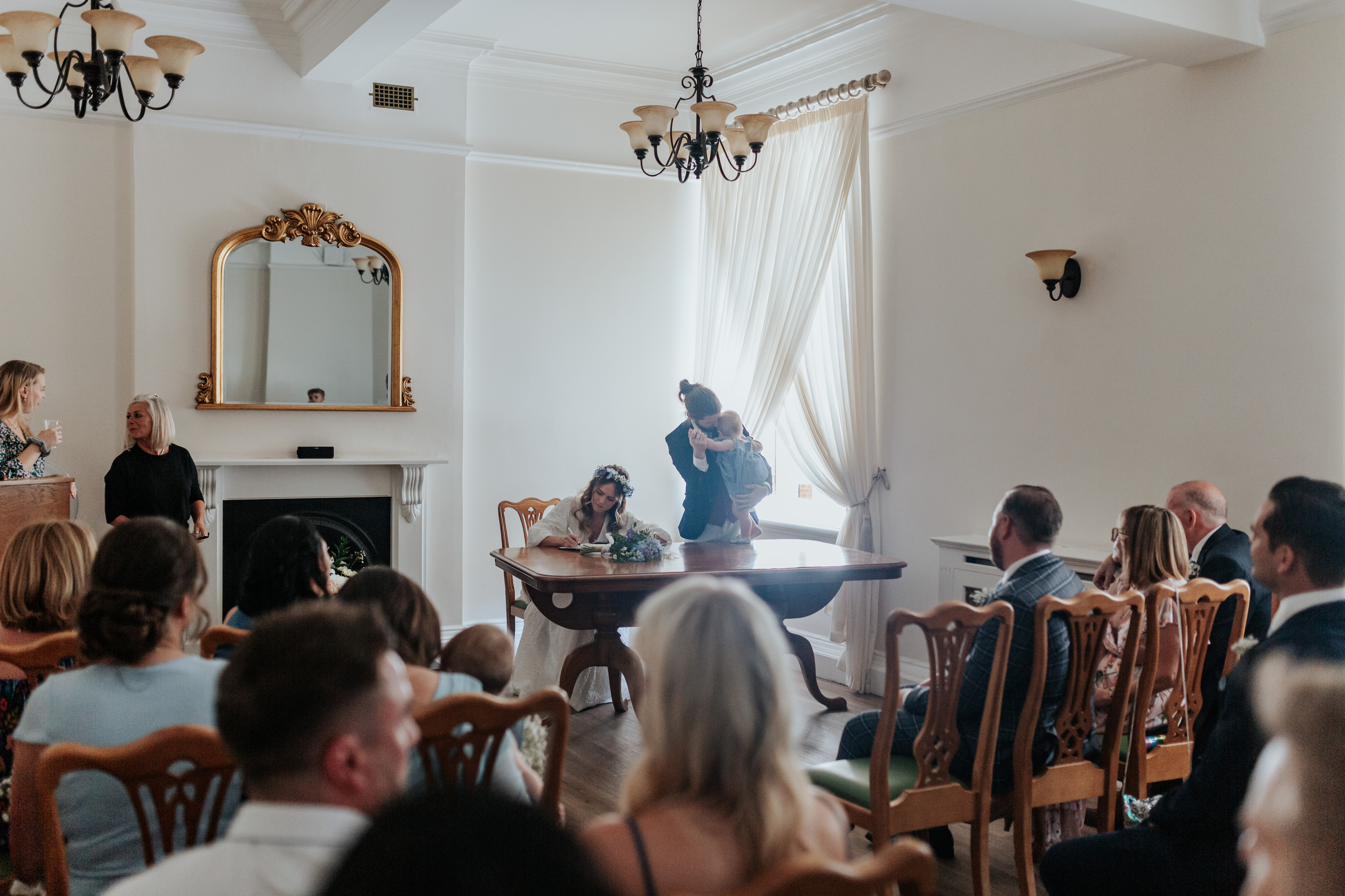 HannahSam25_07-079 Bride and groom sign the register in the Edwardian room of Woolwich Town Hall while the groom dances with his toddler daughter