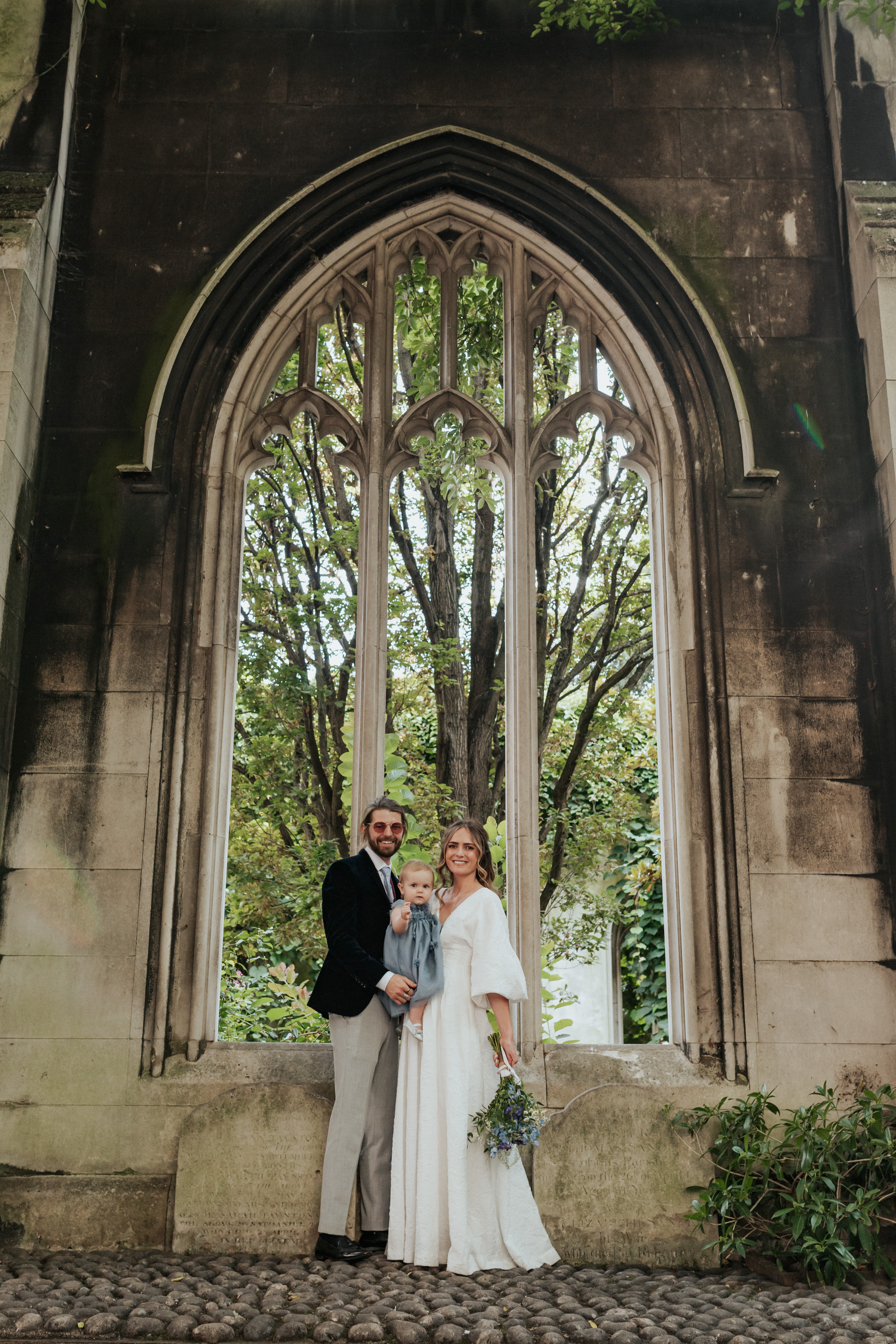 HannahSam25_07-201 Bride, groom and their daughter stood in one of the windows at St Dunstans in The East Gardens