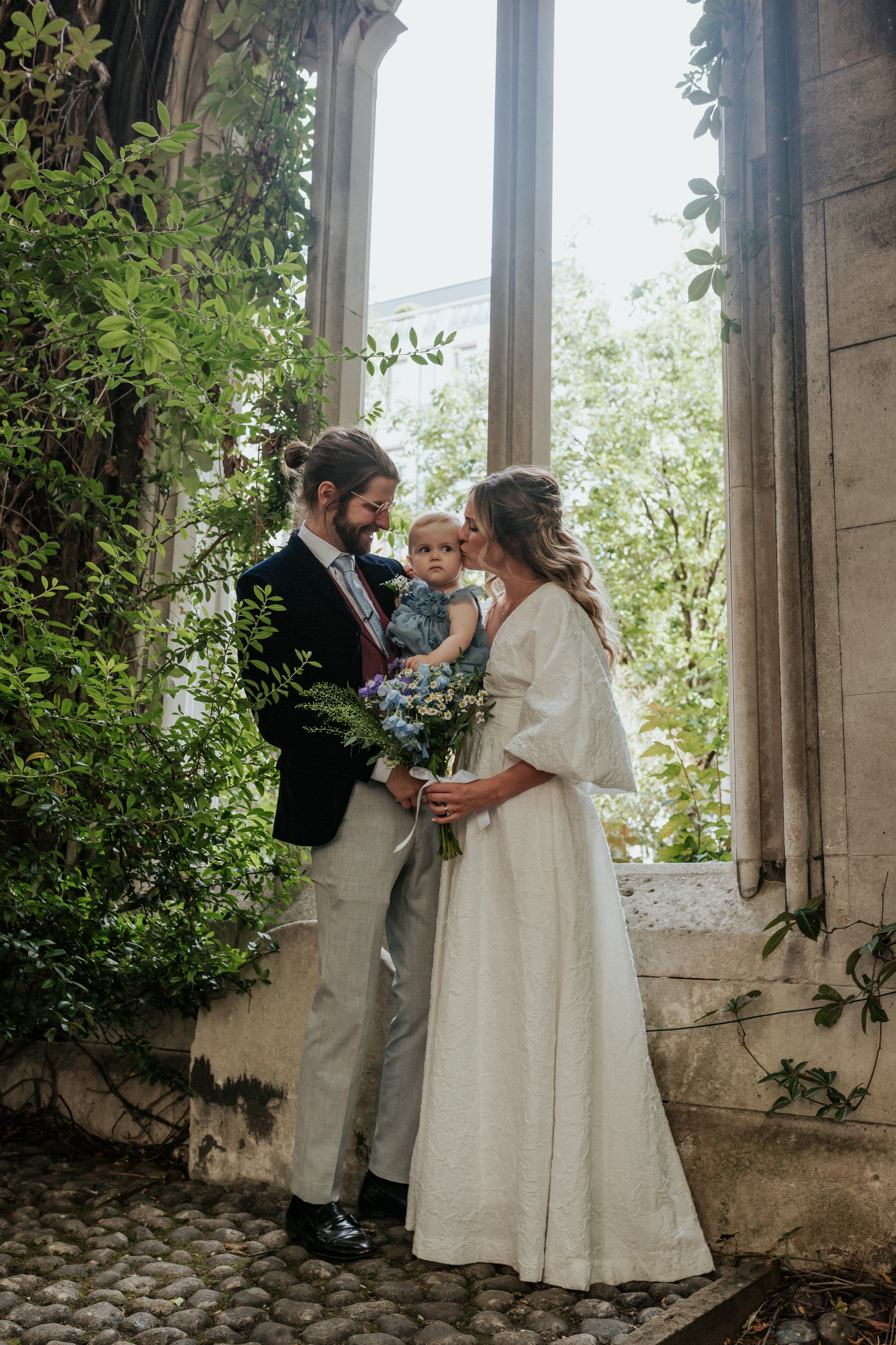 HannahSam25_07-212 Bride, groom and their daughter stood in one of the windows at St Dunstans in The East Gardens
