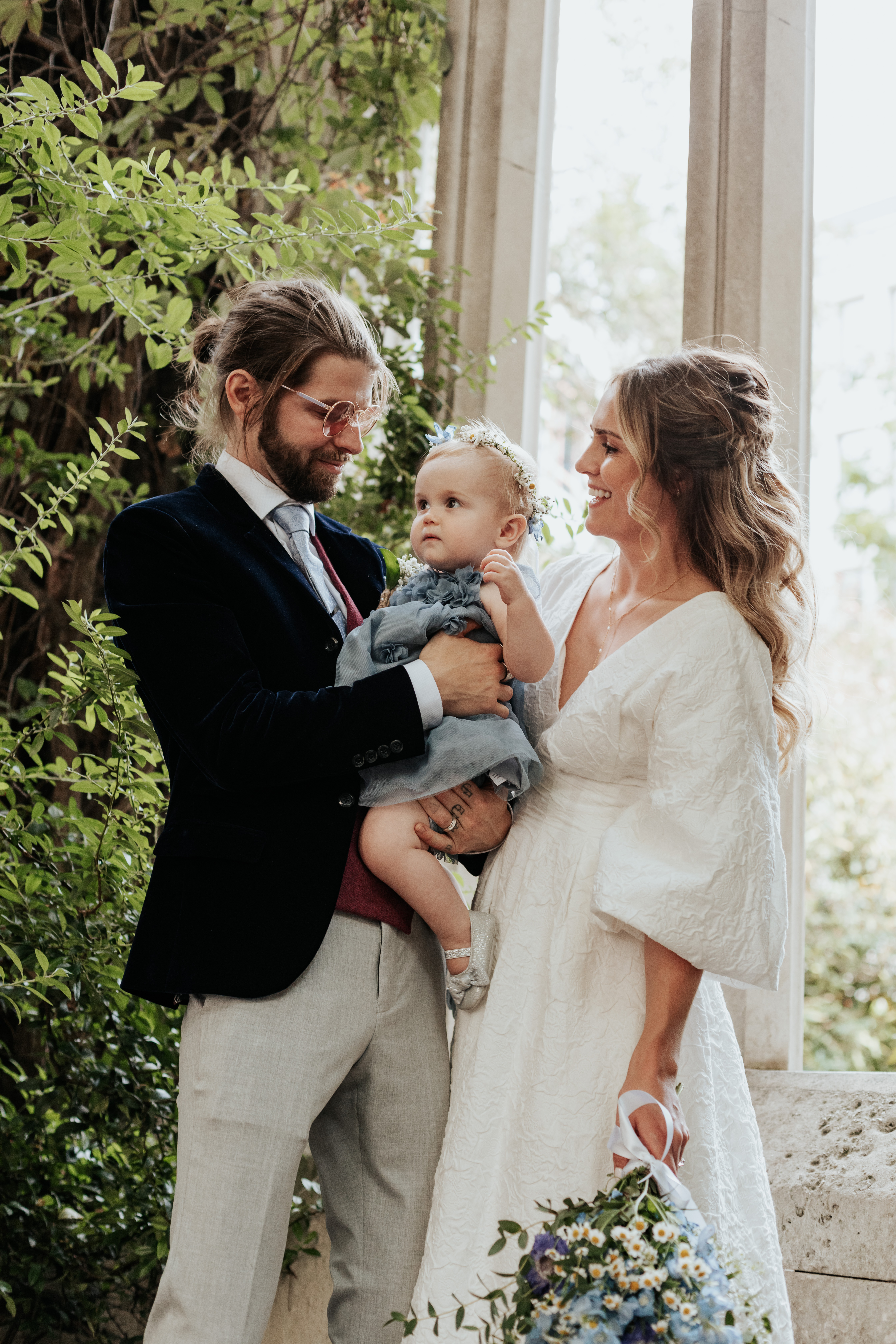 HannahSam25_07-218 Bride, groom and their daughter stood in one of the windows at St Dunstans in The East Gardens