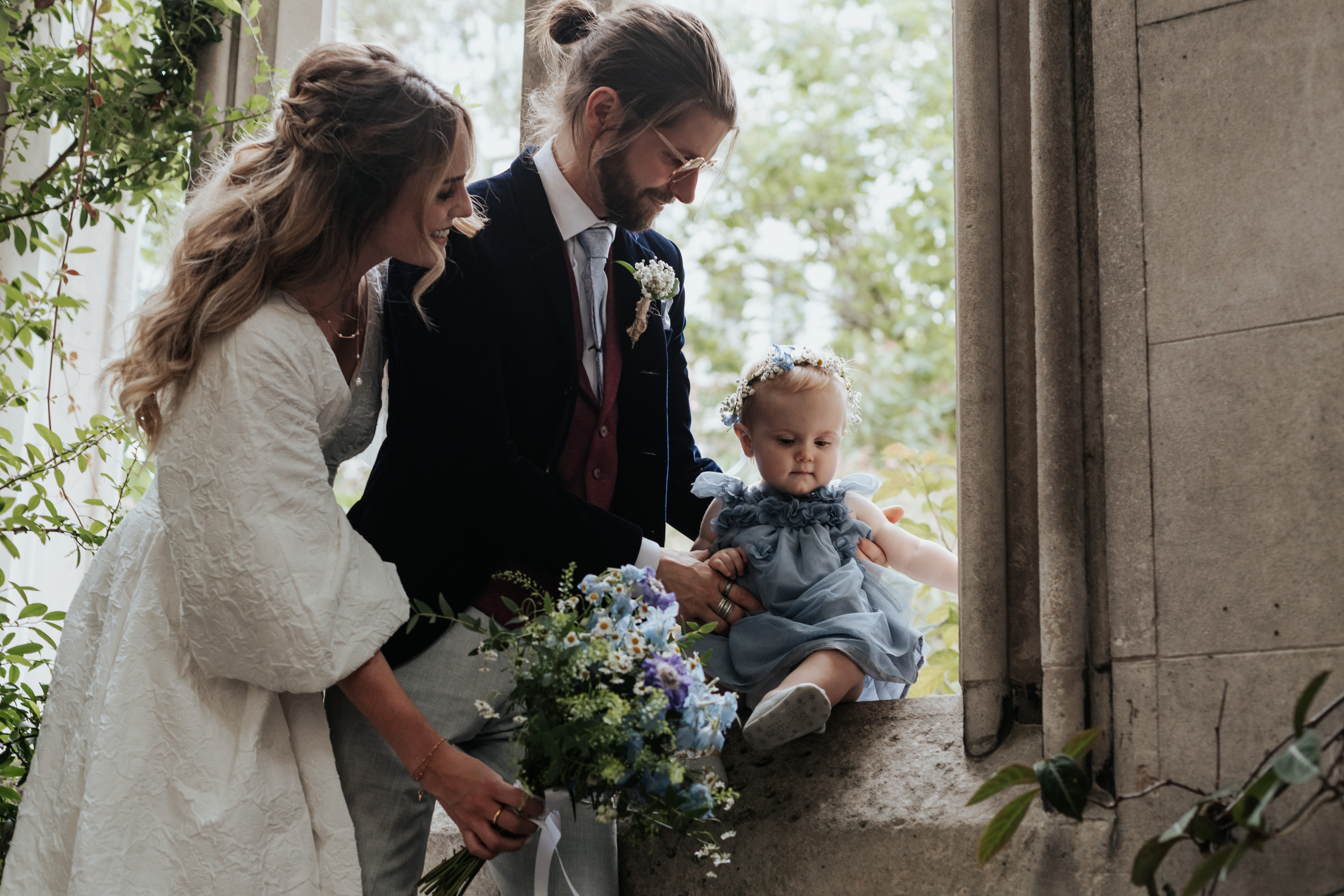 HannahSam25_07-224 Bride, groom and their daughter stood in one of the windows at St Dunstans in The East Gardens