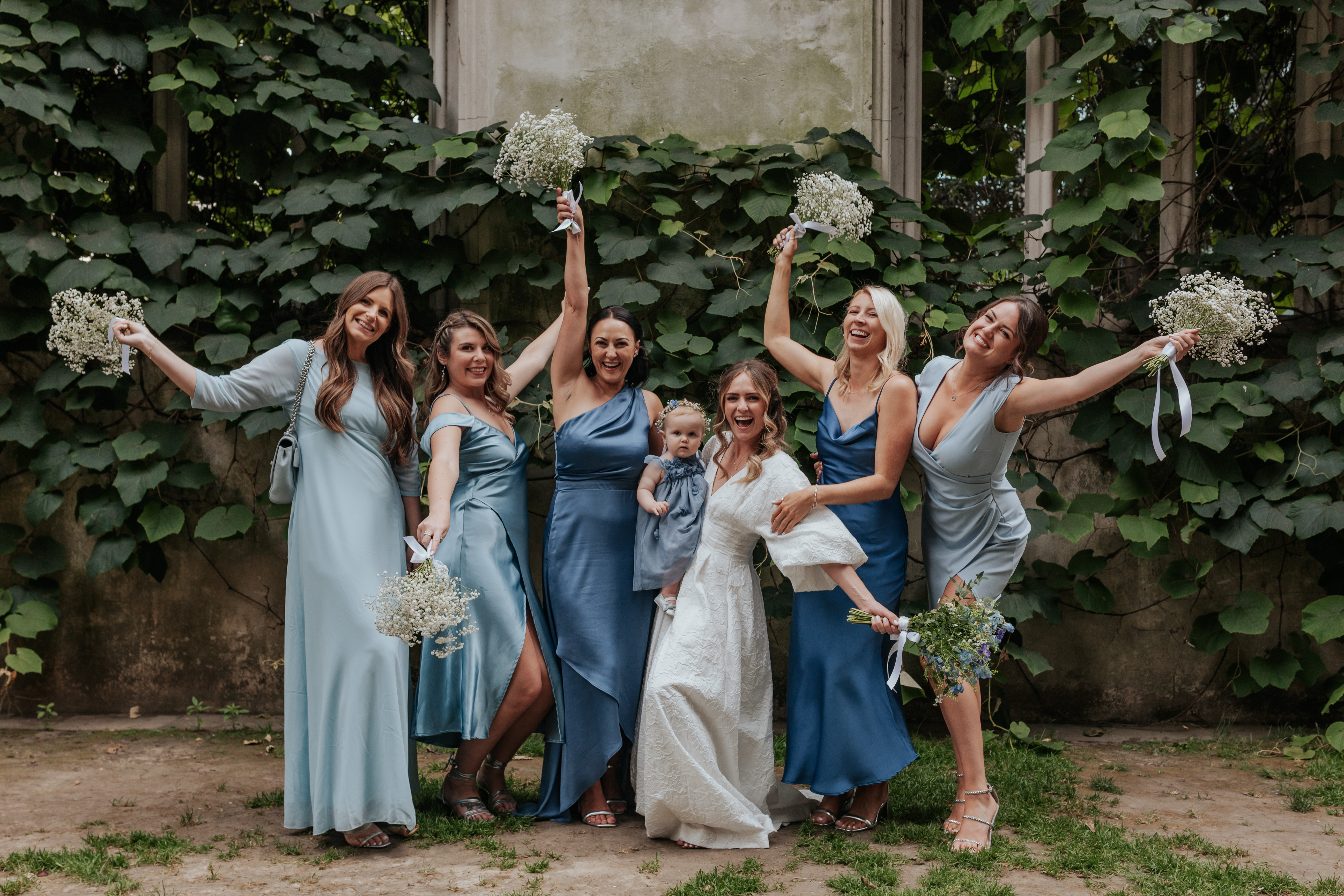 HannahSam25_07-235 Bride and her bridesmaids wearing mismatched blue dresses with their bouquets in the air in St Dunstans in The East gardens