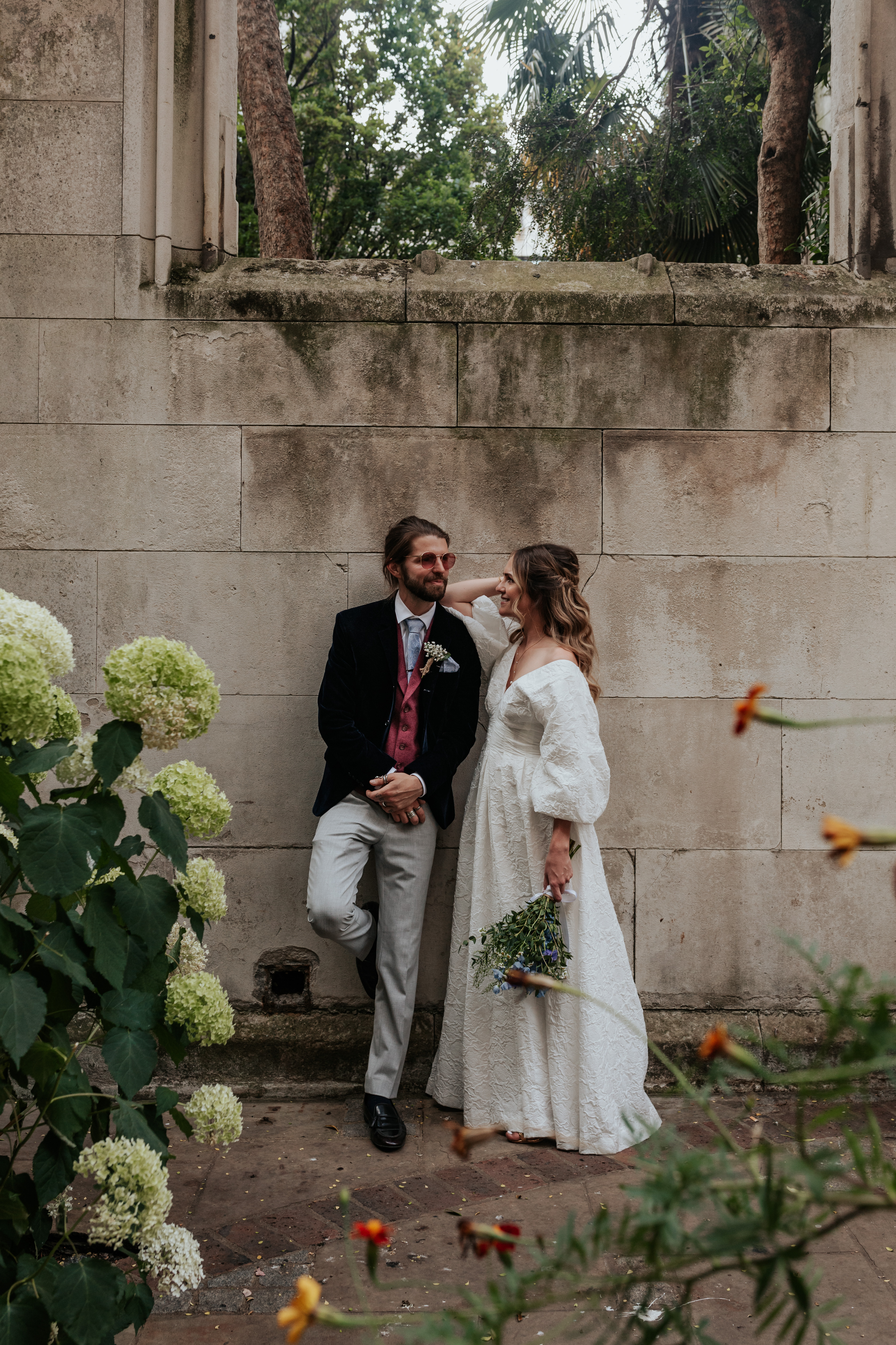 HannahSam25_07-271 Bride and groom lean against a wall atBride and groom stand in one of the windows at St Dunstans in The East gardens during their London Wedding