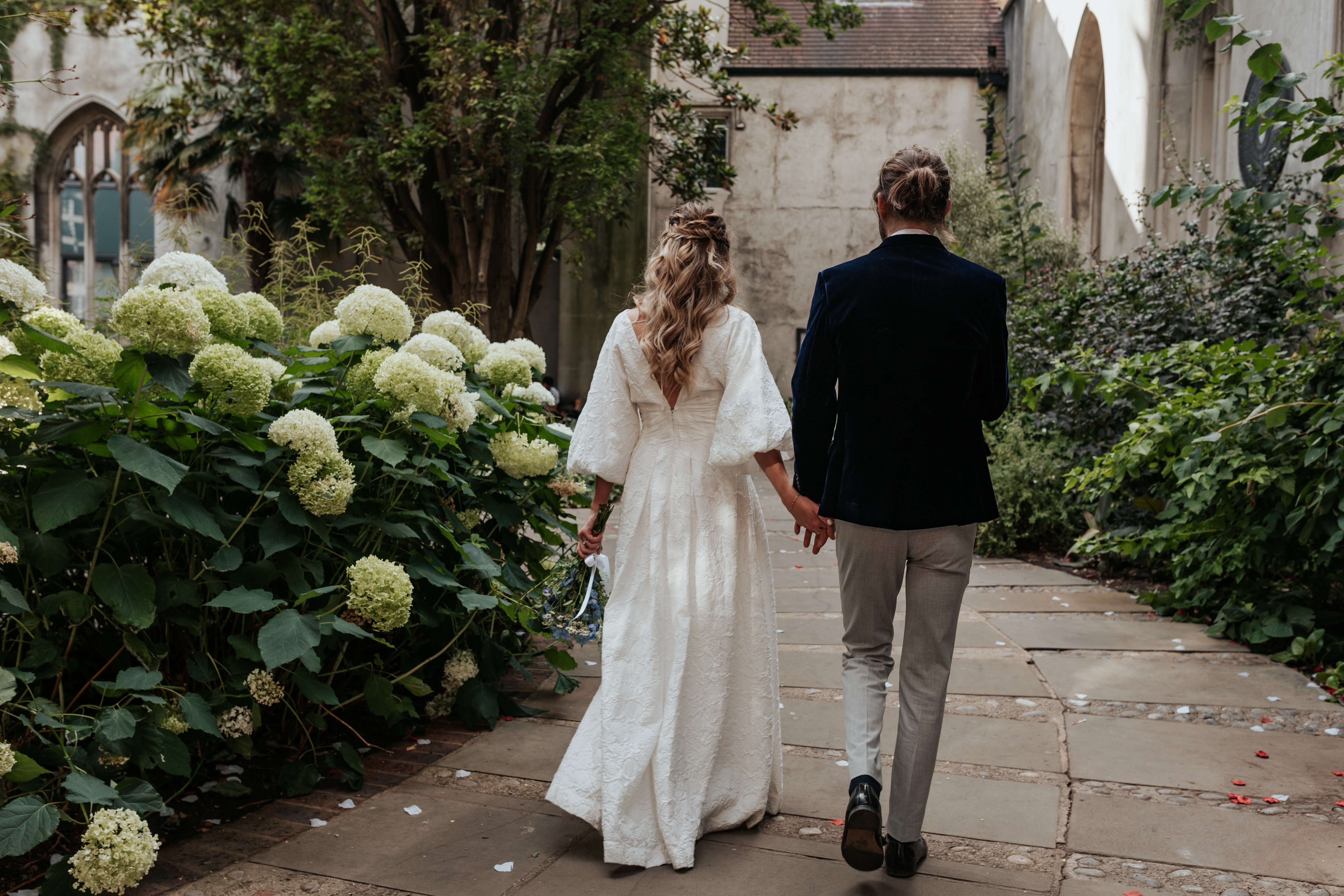 HannahSam25_07-297 Bride and groom walk away holding hands atBride and groom stand in one of the windows at St Dunstans in The East gardens