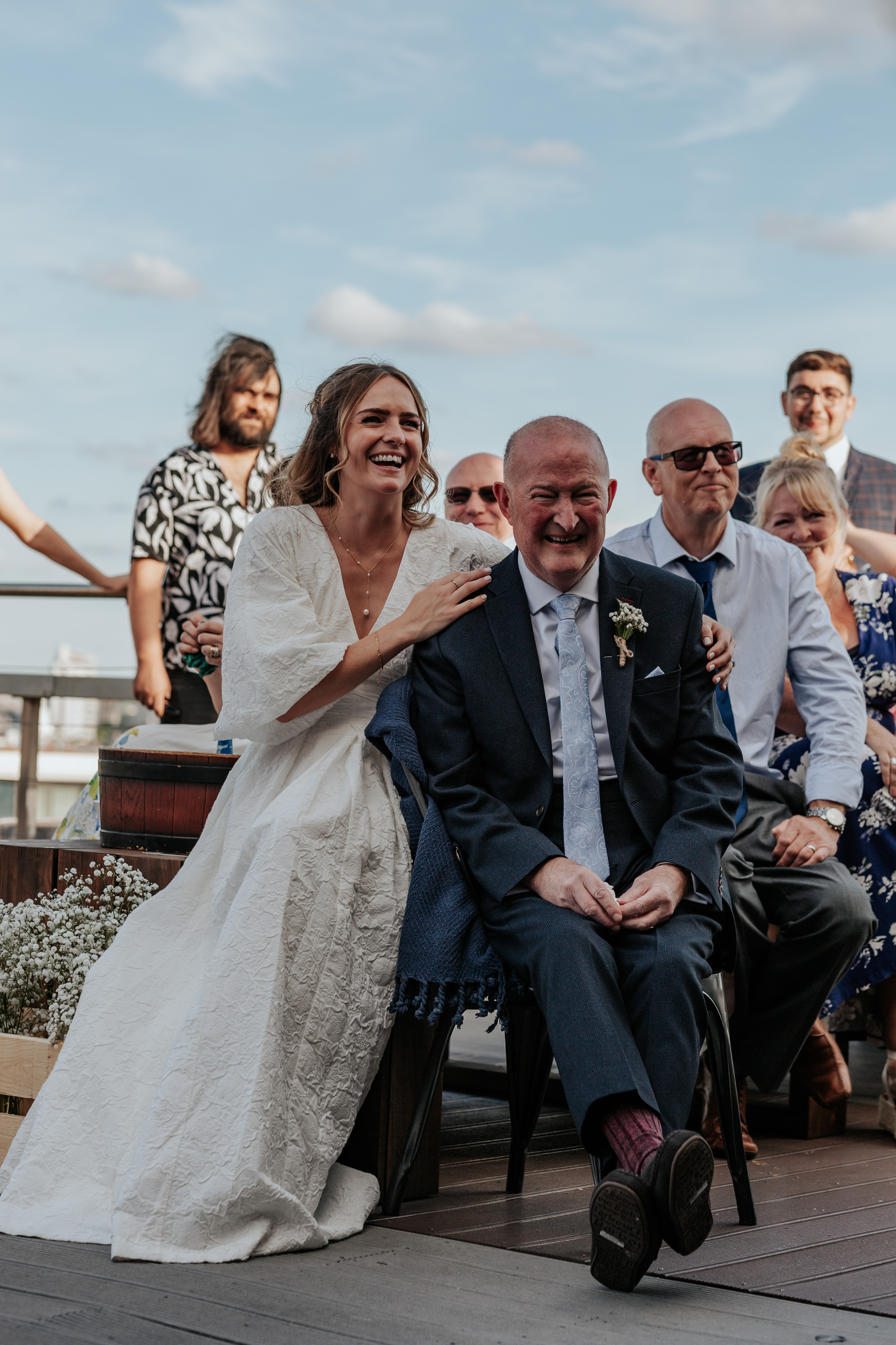 HannahSam25_07-395 Bride sits with her father in law and laughs during the speeches on a London rooftop terrace