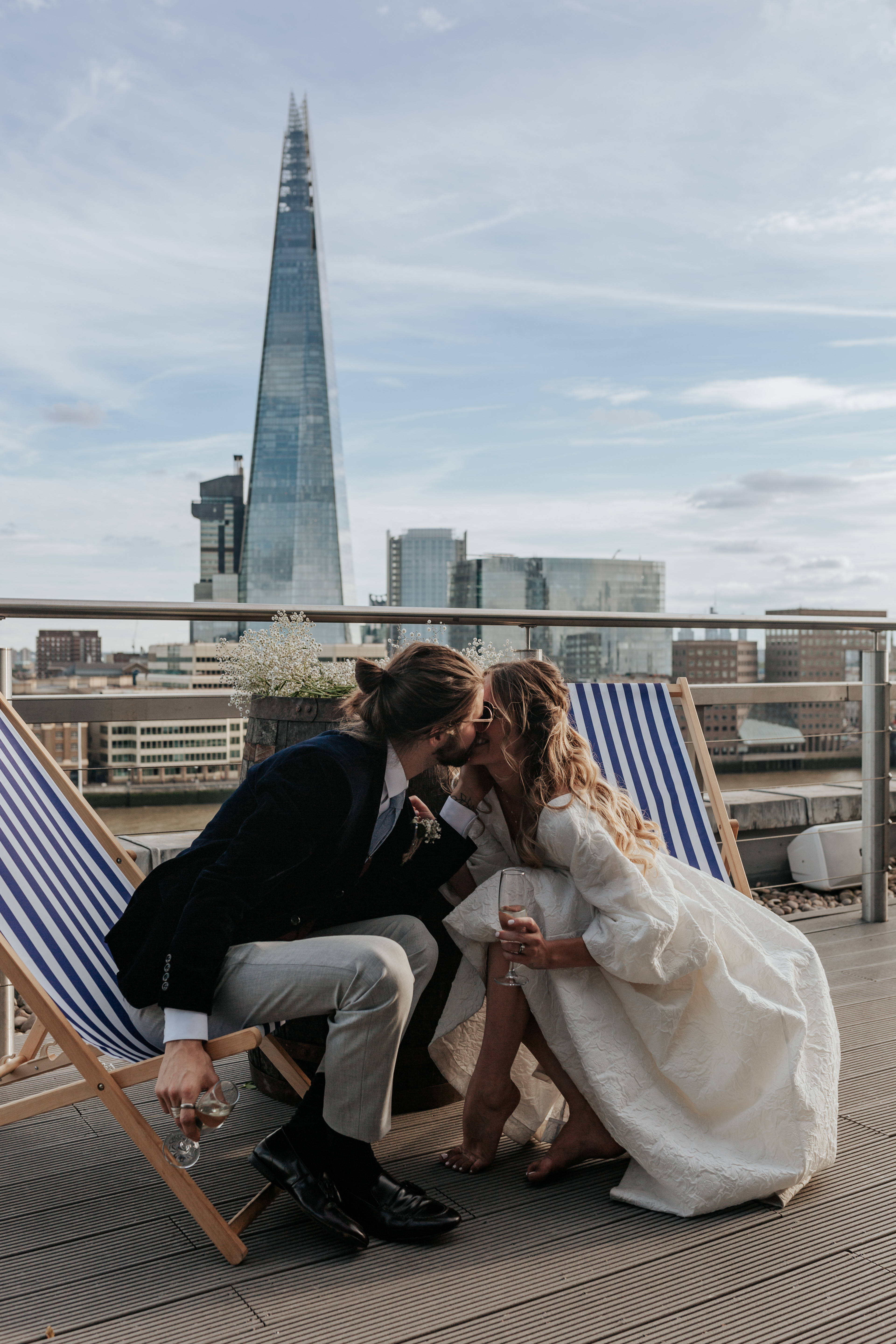 HannahSam25_07-453 Bride and groom kiss in deck chairs on the rooftop terrace of a London bar with The Shard in the background