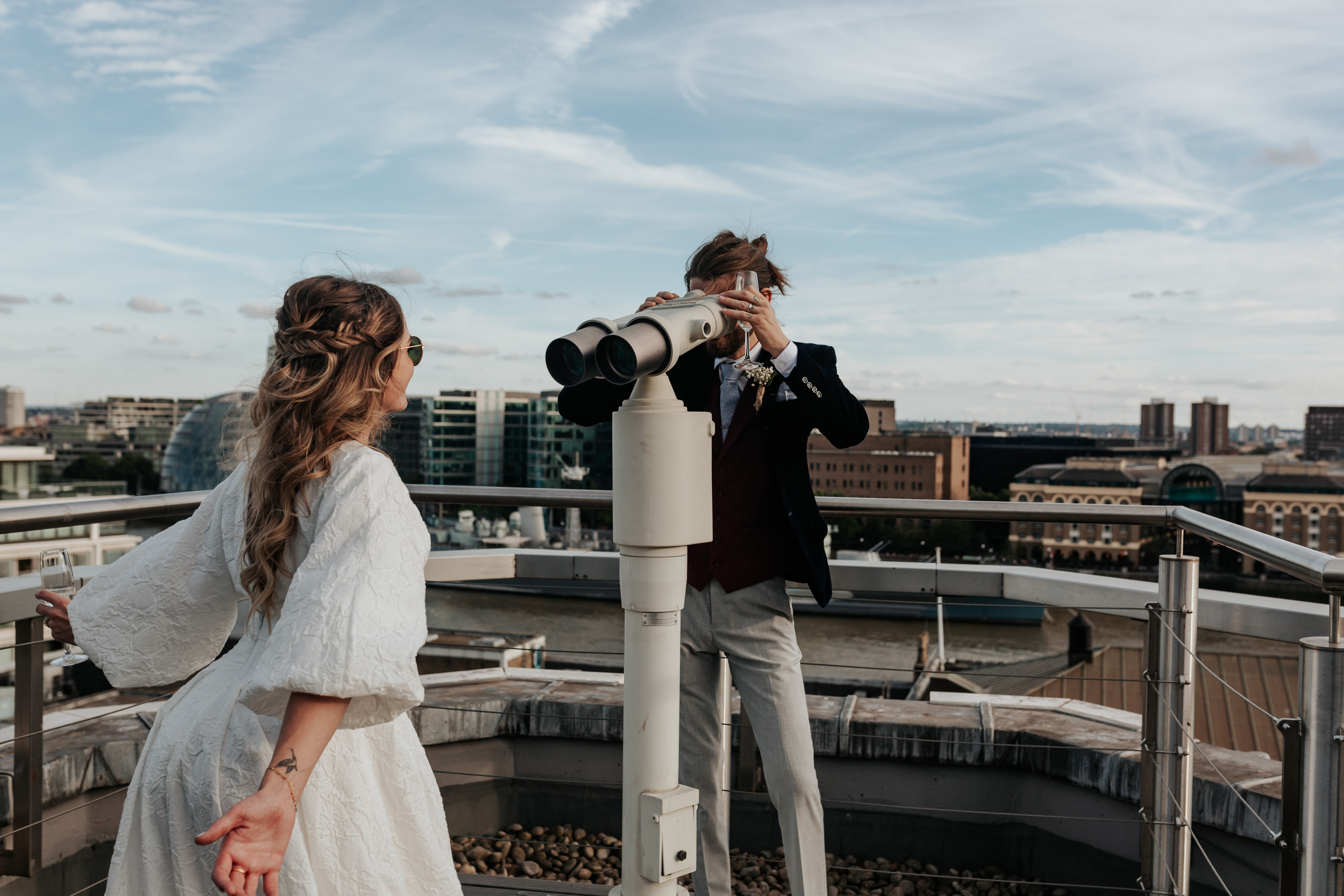 HannahSam25_07-459 Groom looks at the bride through viewfinder binoculars on the rooftop terrace of a London bar