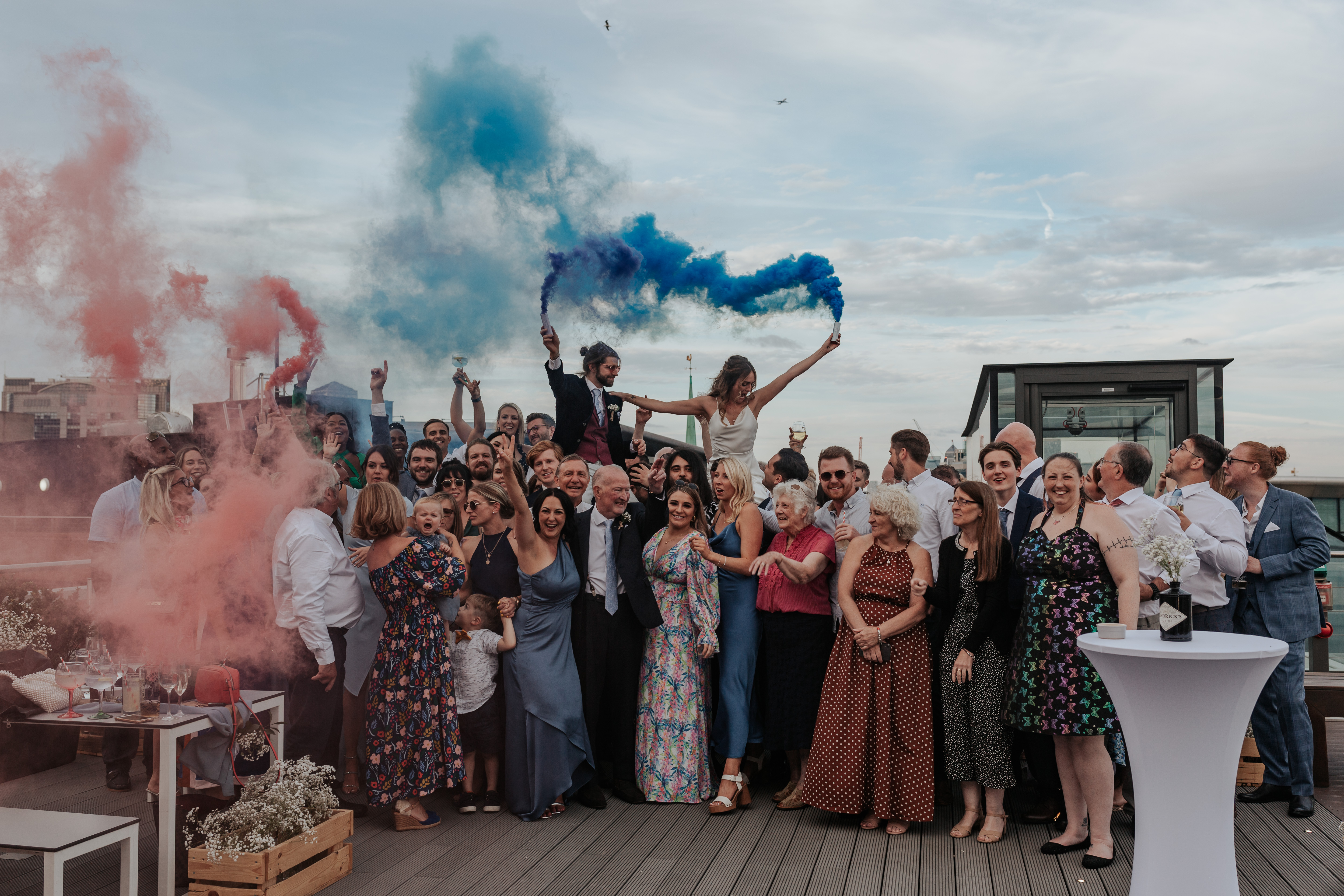 HannahSam25_07-577 Bride and groom sit on their guests shoulders and let off smoke flares on the rooftop terrace of a London bar