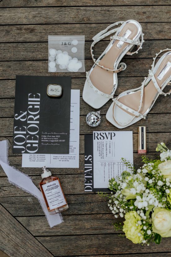 A flatlay of minimal black and white wedding details featuring shoes, stationery, jewellery and flowers