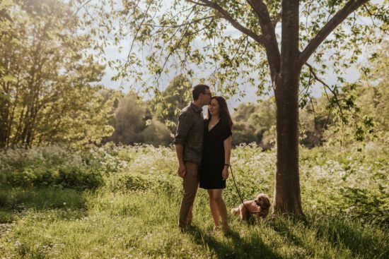 A golden hour photo of couple kissing during their engagement shoot while their dog looks at them