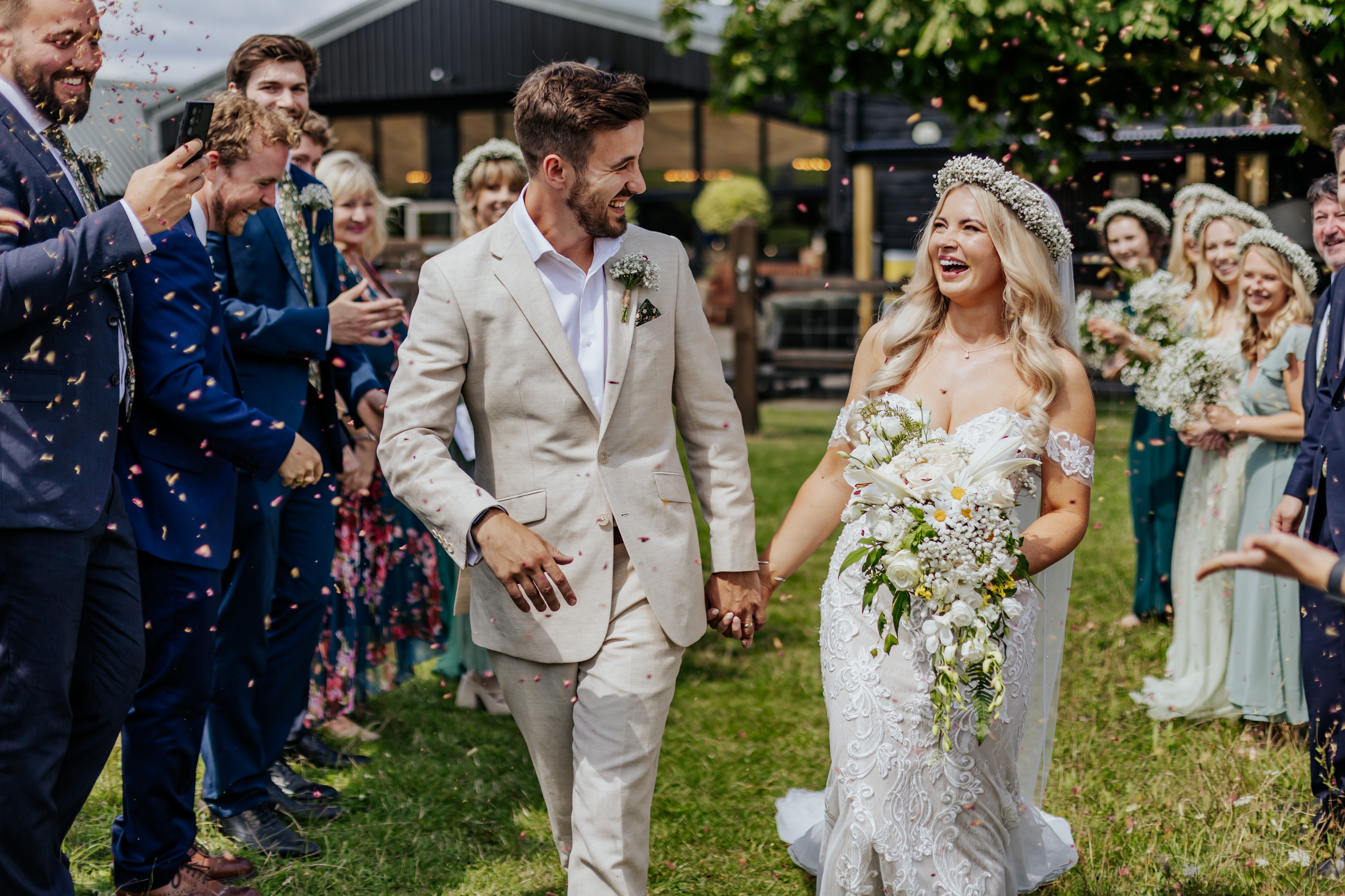 Bride and groom walk through a confetti tunnel with Wellington Barn in the background