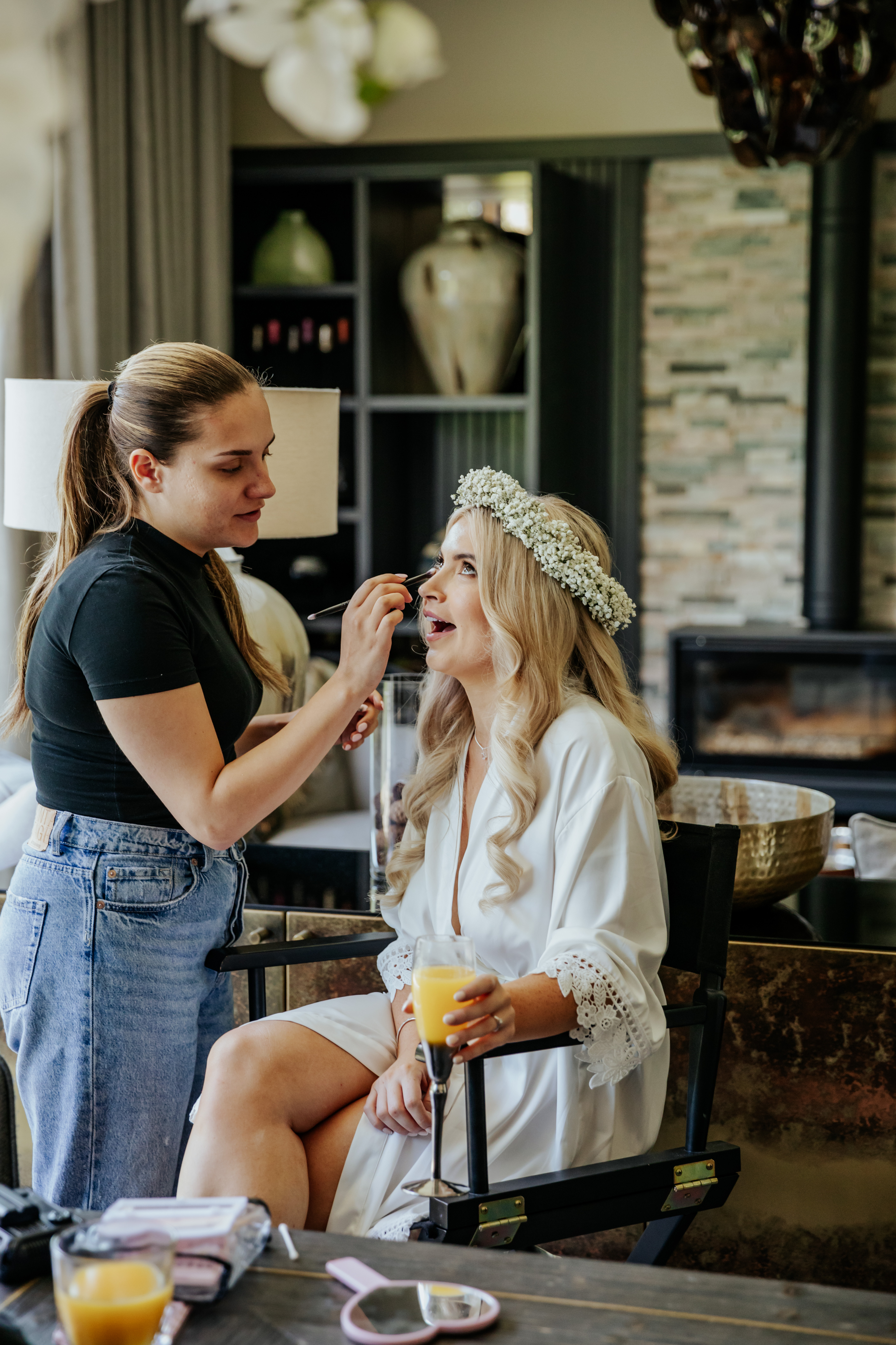 Bride Frankie in a white dressing gown and flower crown laughs as she gets her makeup done