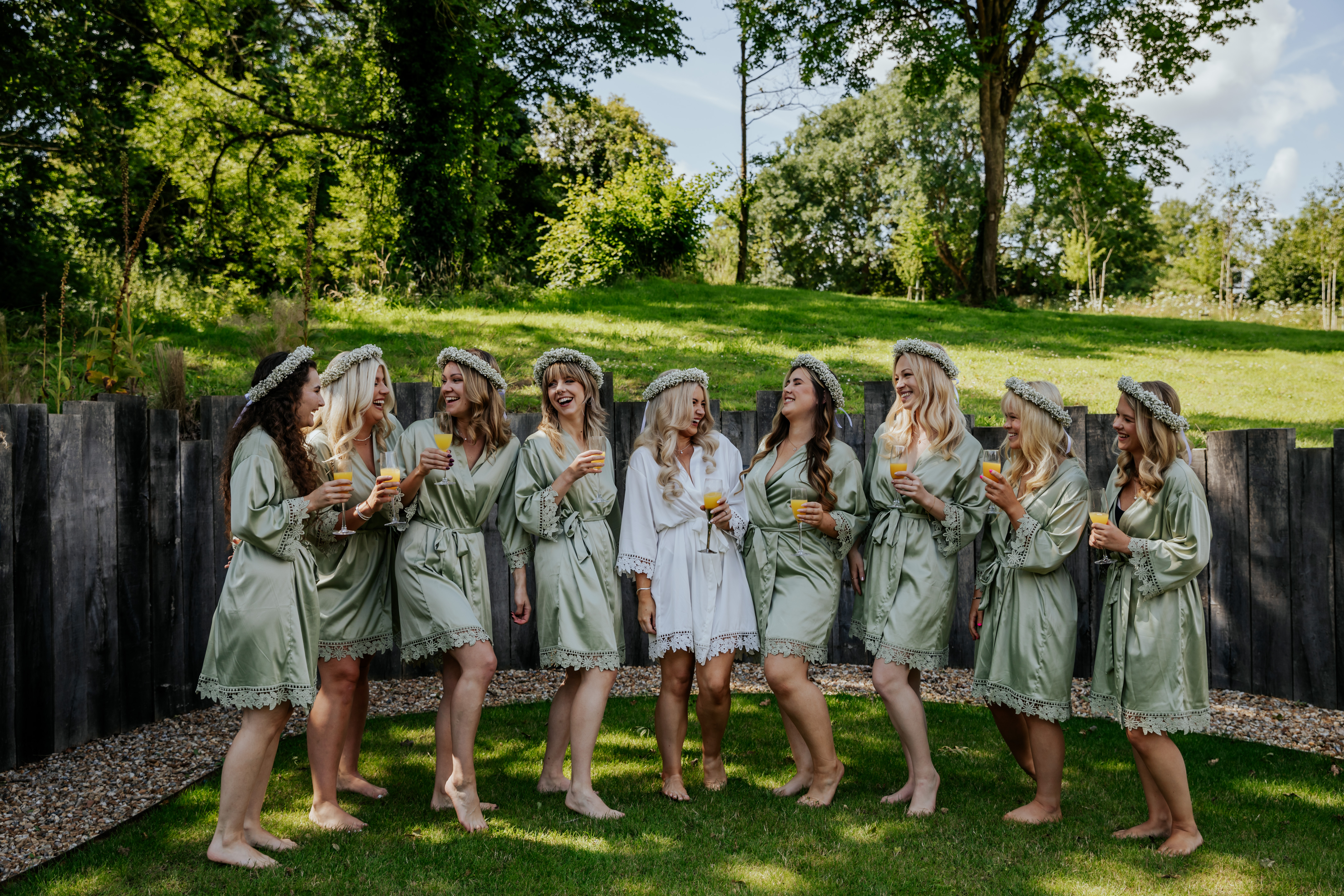 Bridal party laugh together in matching dressing gowns and flower crowns, holding mimosas