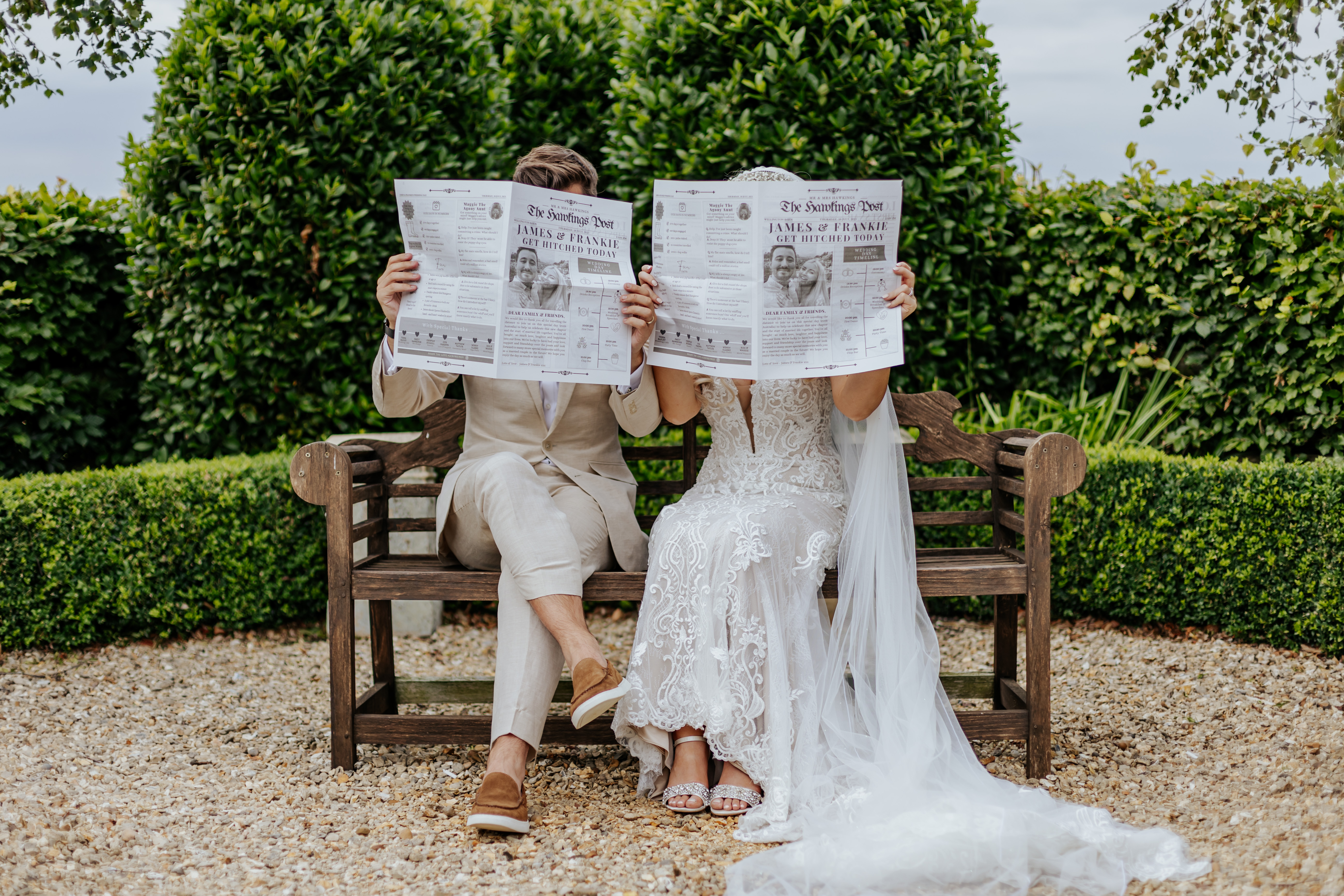 Bride and groom sat together on a bench, hiding their faces with their wedding newspapers outside Wellington Barn