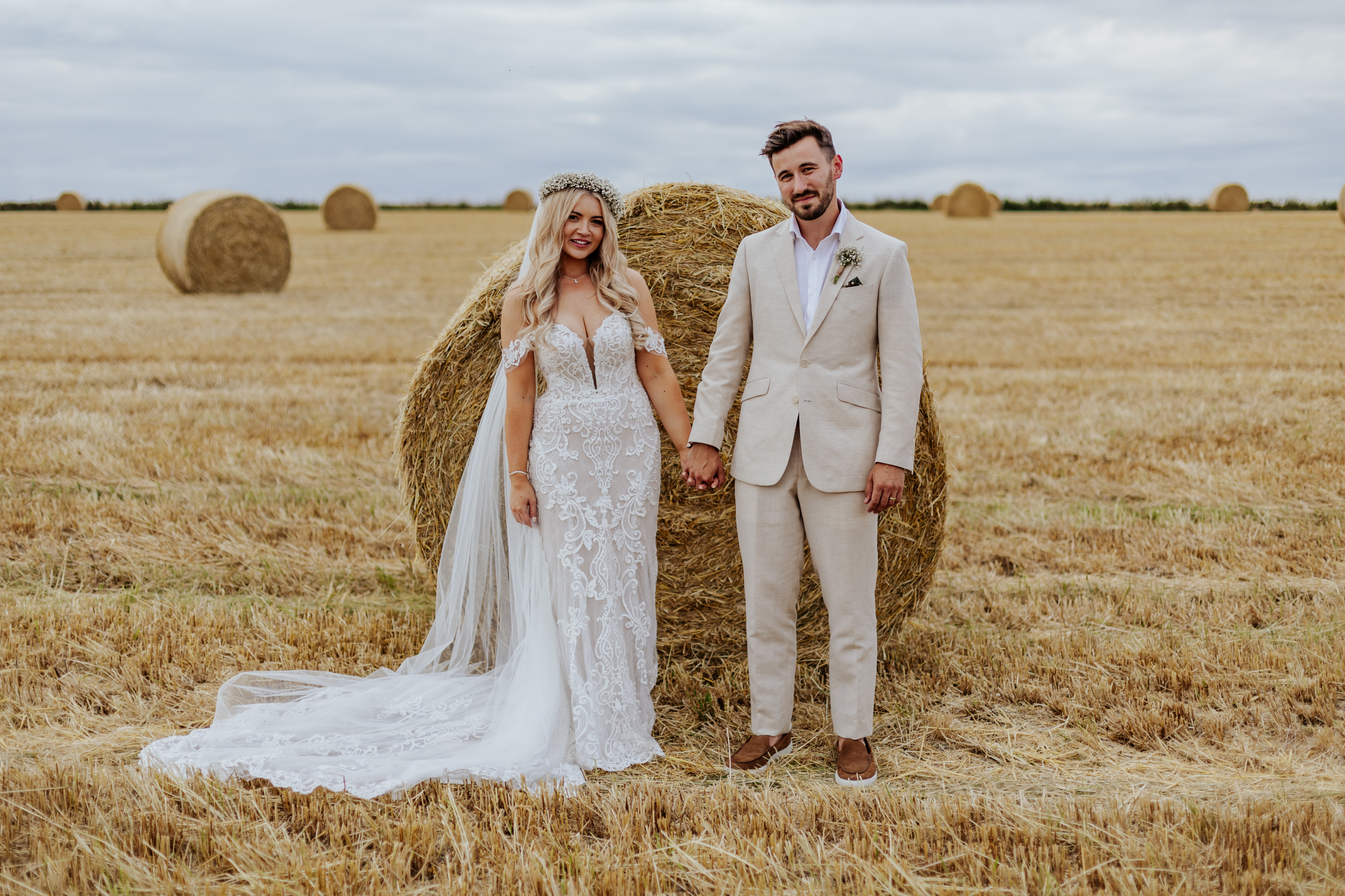 Bride and groom hold hands and look at the camera infront of a circular hay bale