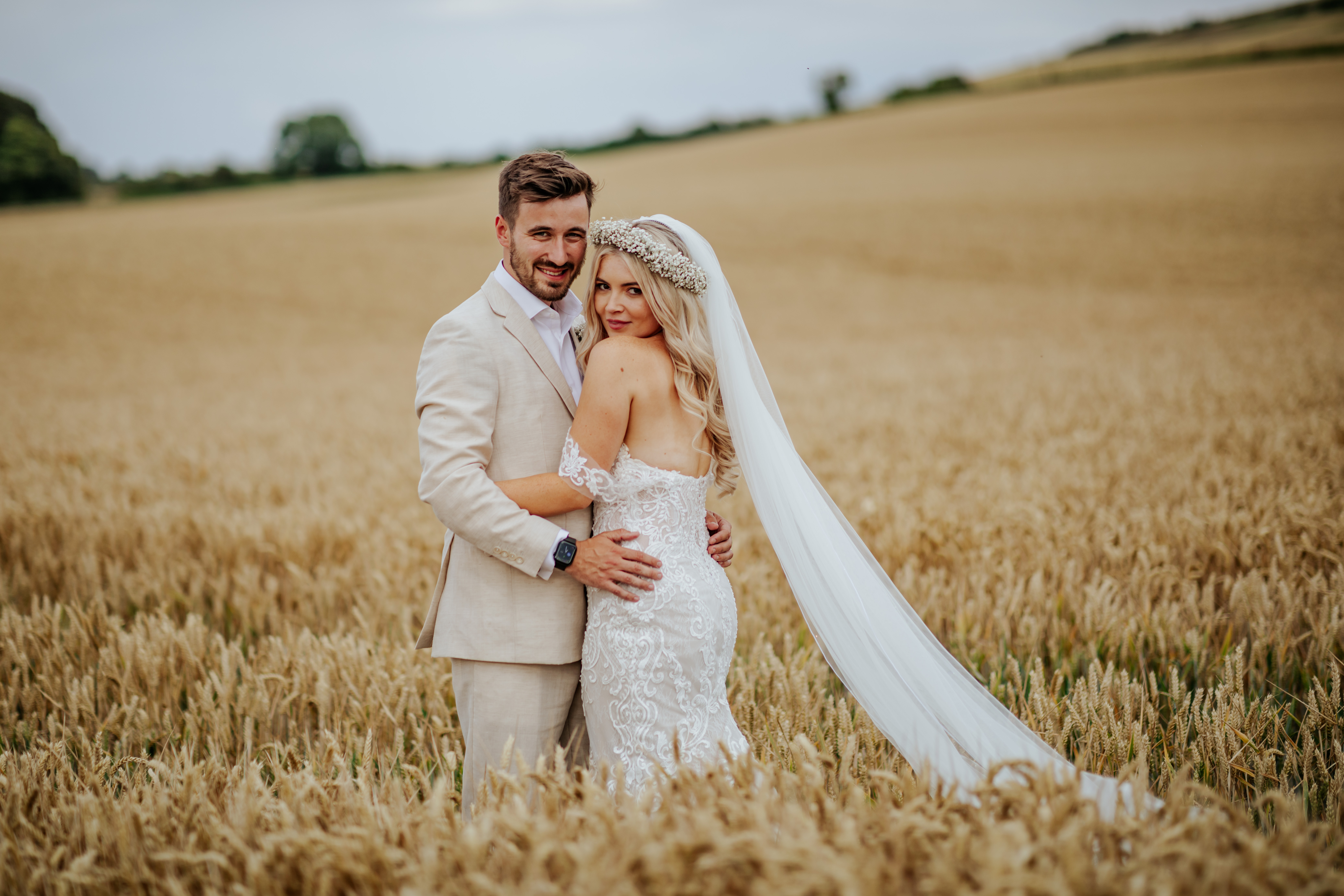 Bride and groom stand in a field of wheat. Bride looks over her shoulder at the camera